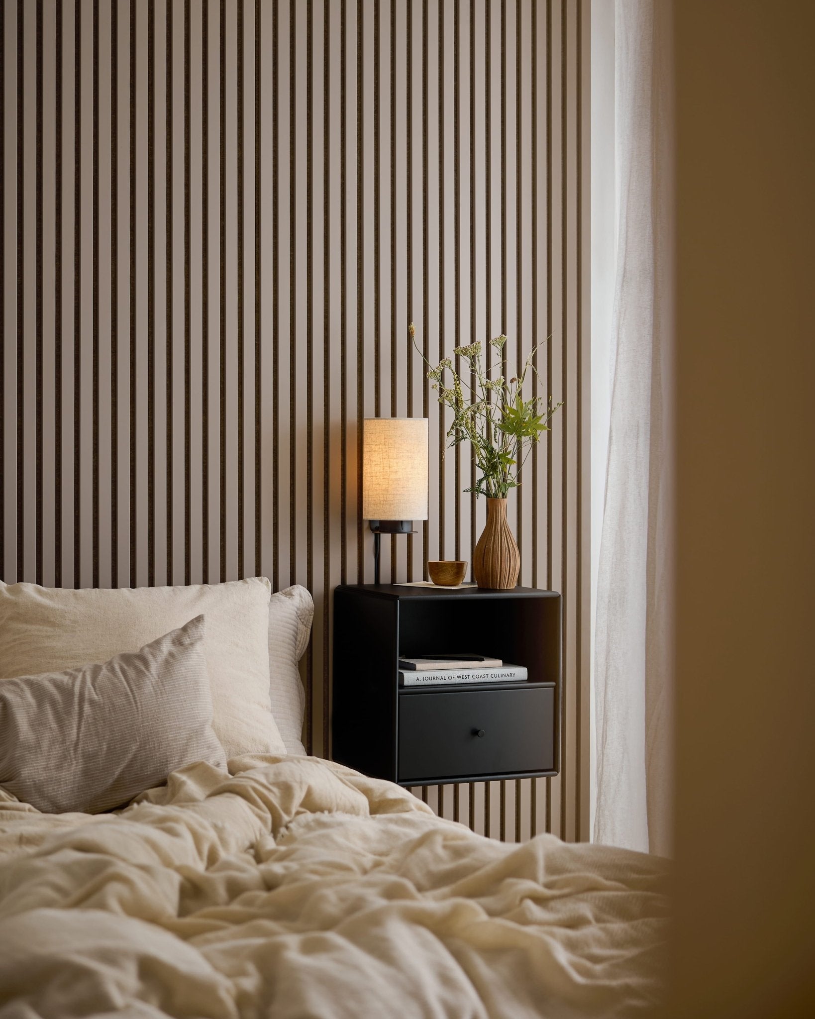 En seng med cremefarvet sengetøj ved siden af et sort natbord foran en Akupanel-væg i Dark Cashmere.