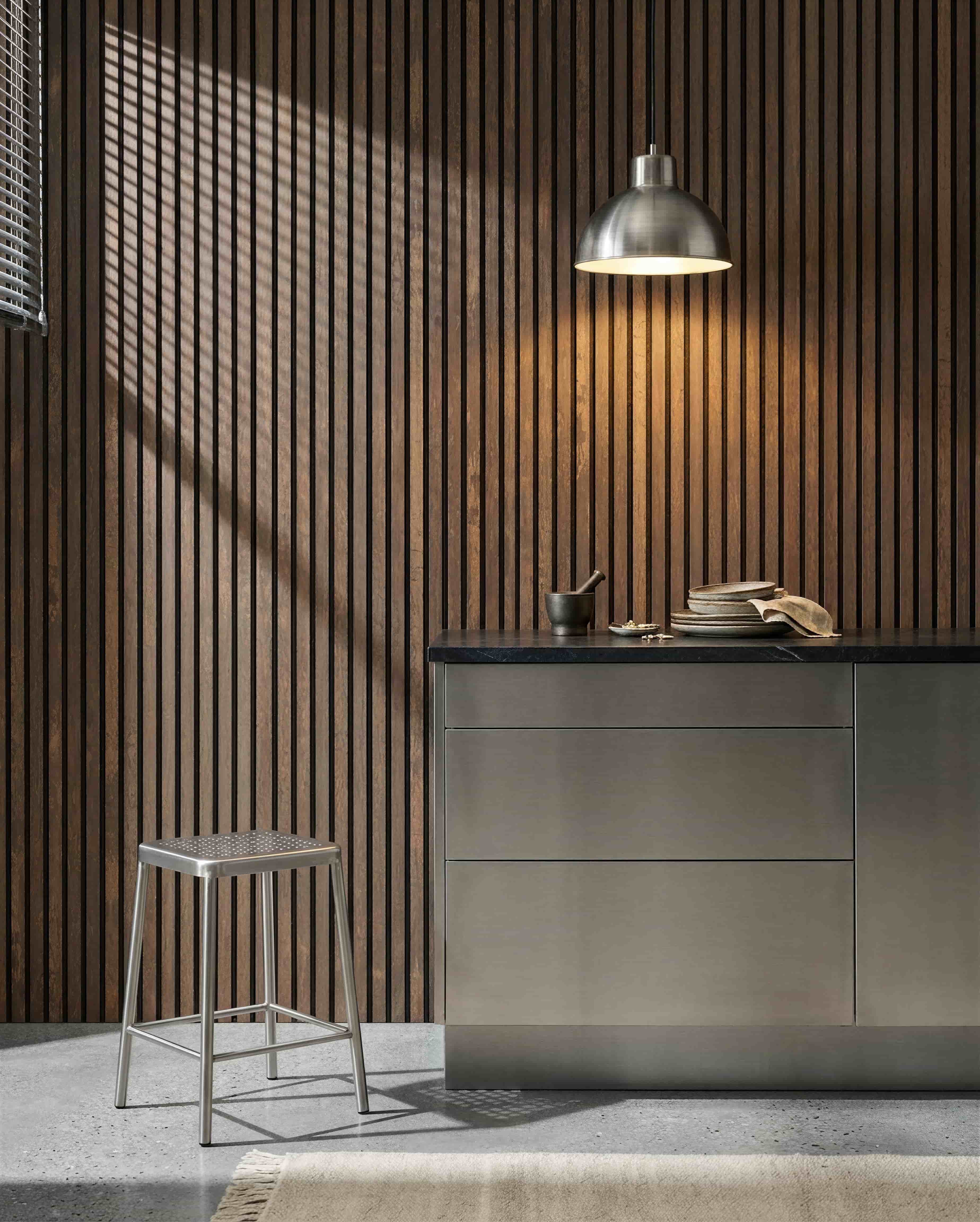Køkken med en WoodUpp Akupanel Oxide Copper akustisk lamelvæg, bordplade i rustfrit stål, metalstol, pendellampe, forskellige tallerkener samt morter og støder.