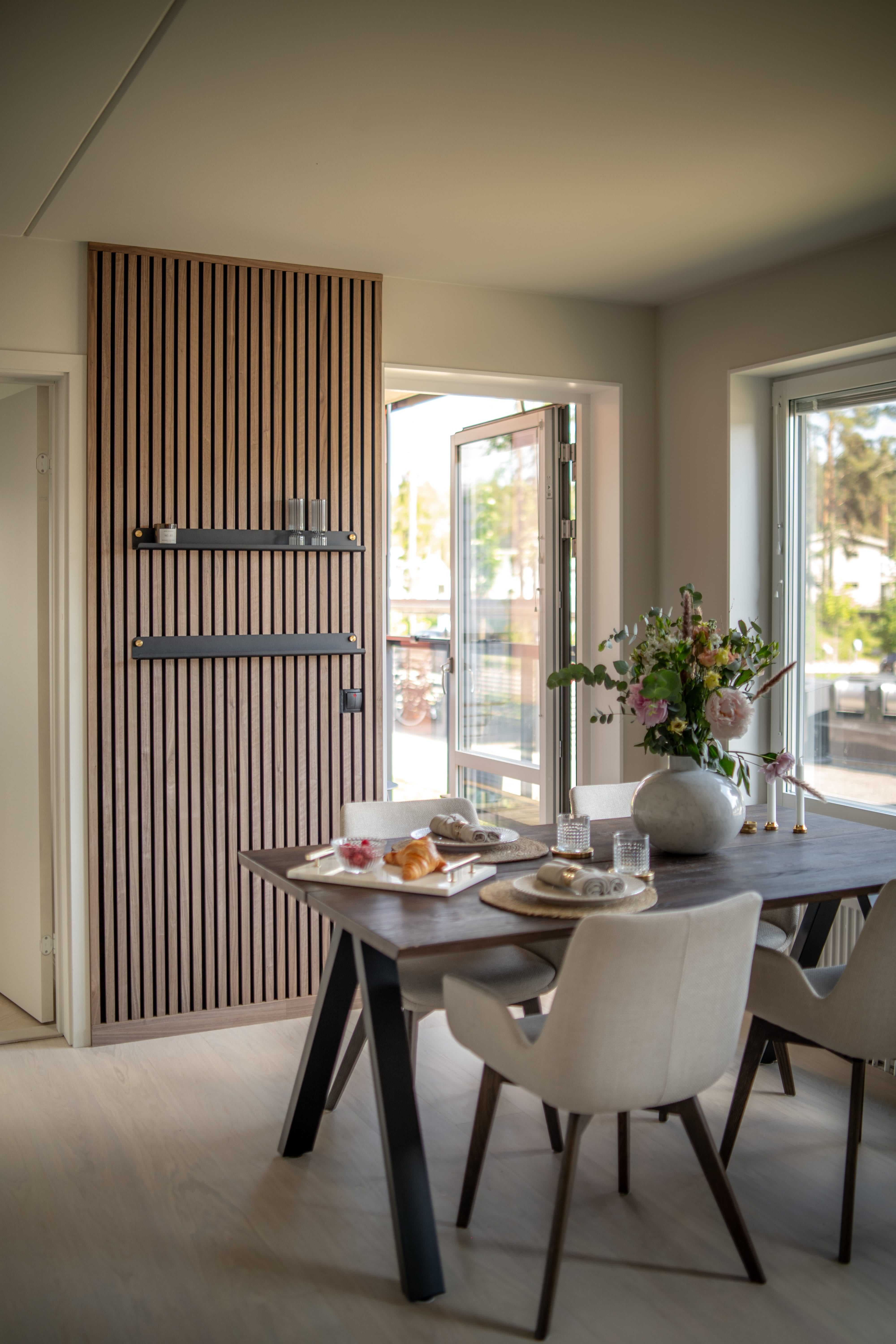 Moderne spisestue med Akupanel akustisk lamelvæg, mørkt morgenbord, minimalistiske beige stole og en vase med blomster ved store åbne vinduer.