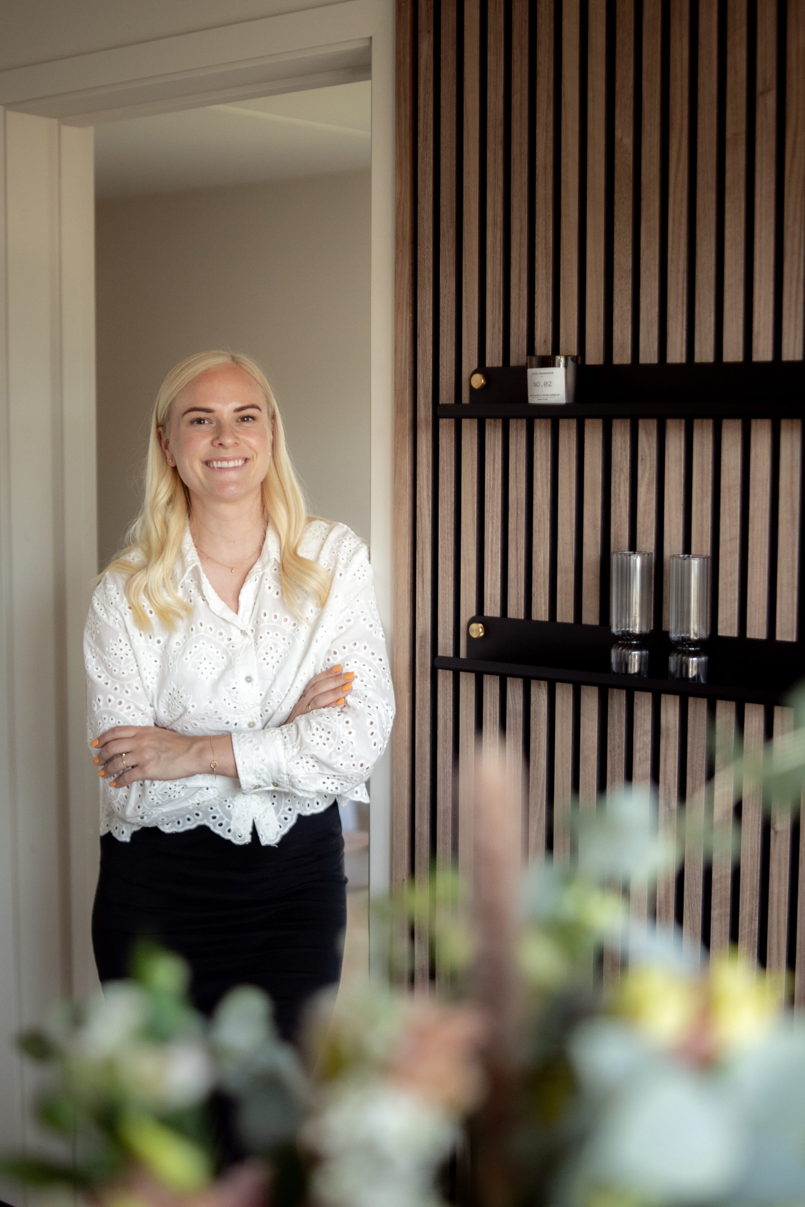 En kvinde med langt blondt hår og hvid bluse står smilende med armene over kors i et moderne rum med nordiske akustiske træpaneler og slanke hylder.