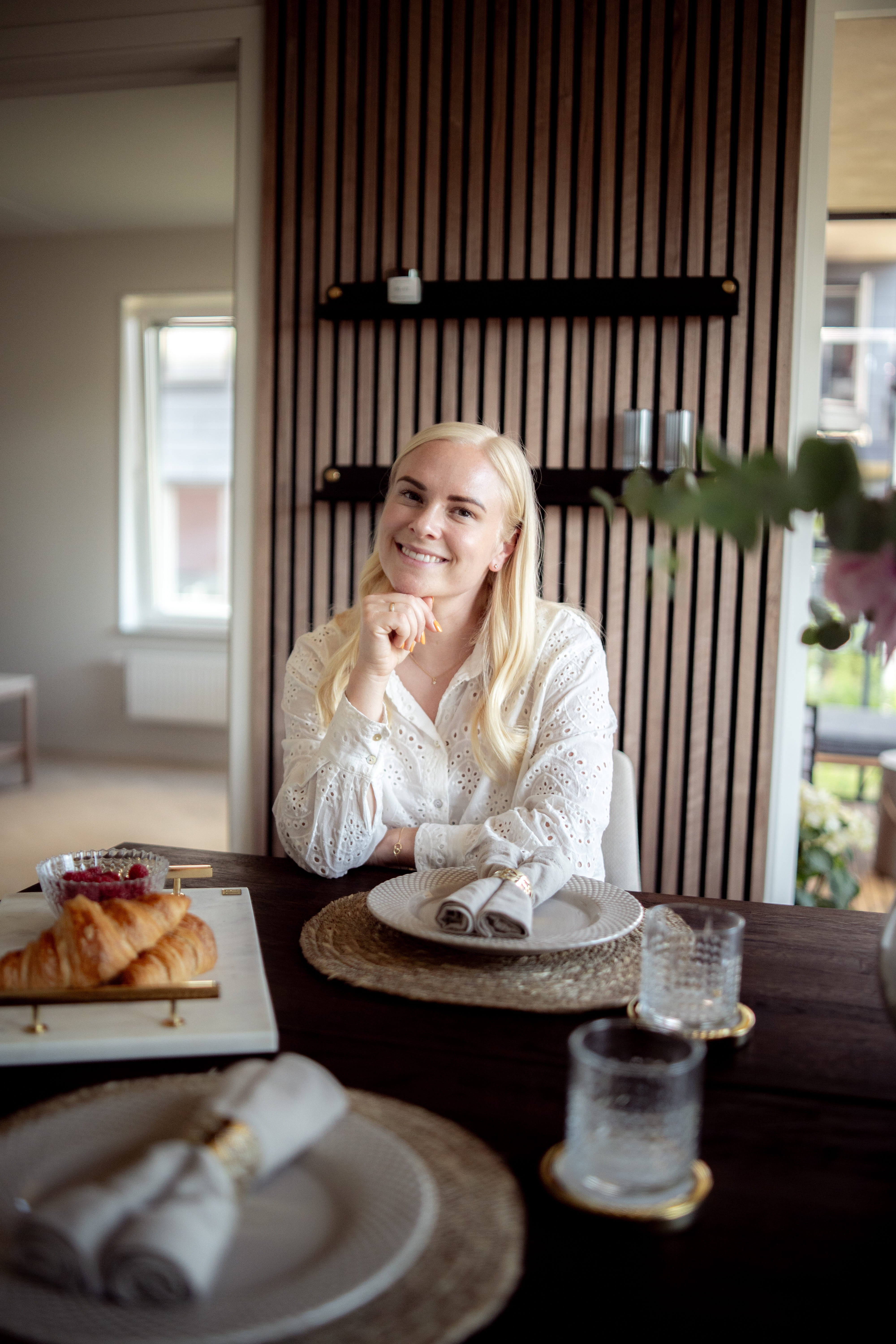 En kvinde med langt blondt hår sidder ved et spisebord dækket til morgenmad med croissanter og glas ved siden af et nordisk akustikpanel på væggen.