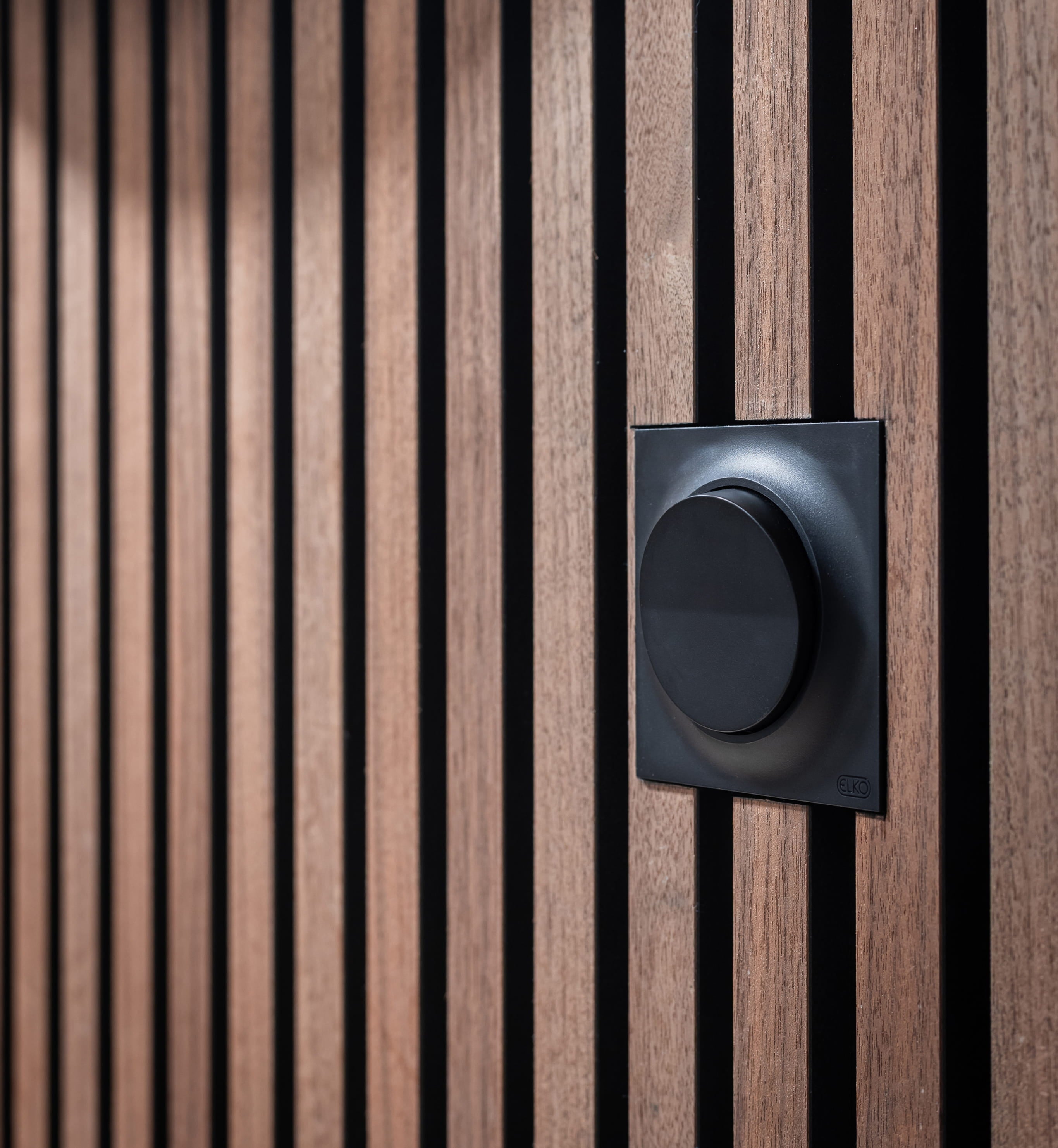 A black circular switch is mounted on a WoodUpp Akupanel wall with vertical wooden slats and deep black gaps between each panel.