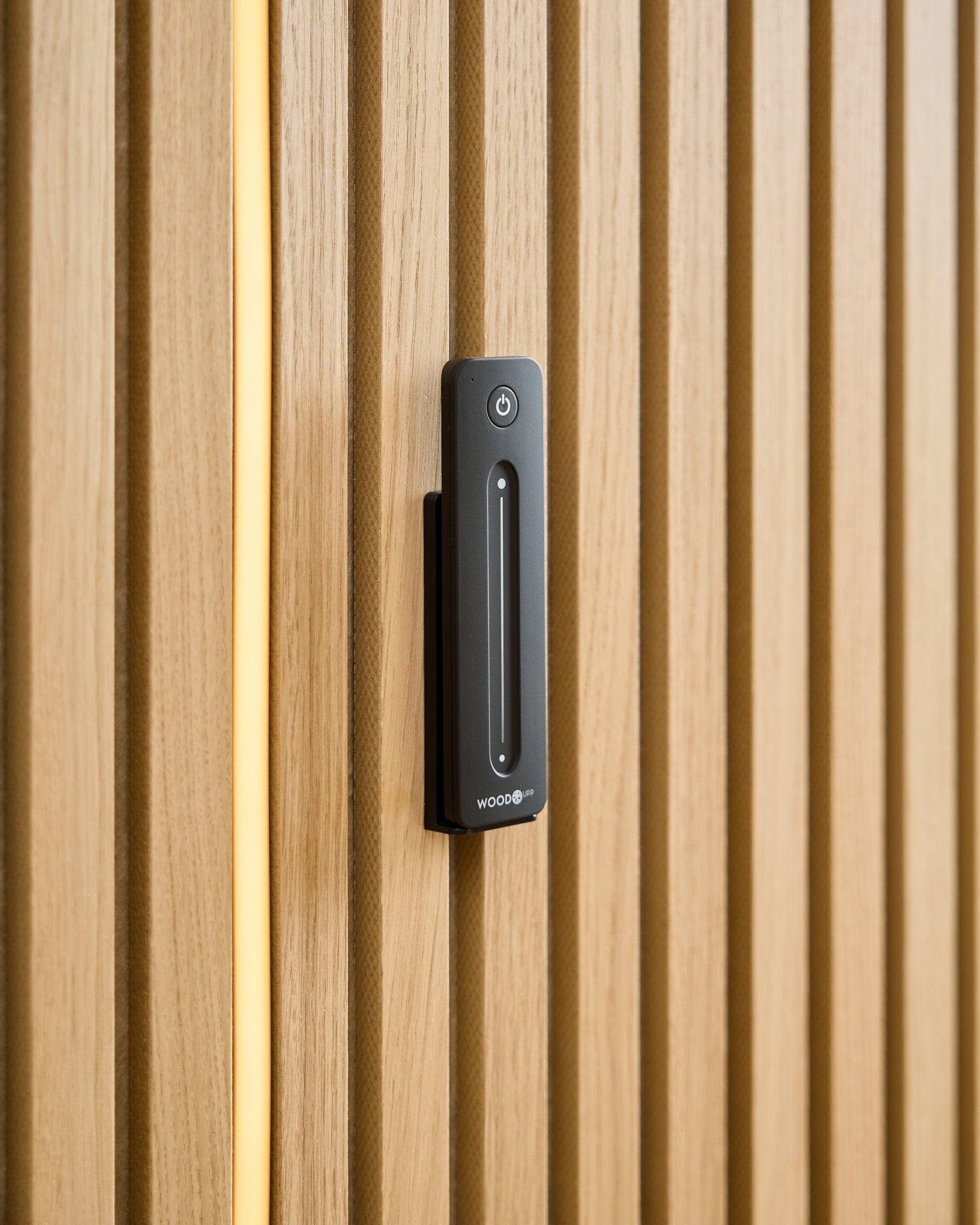 En WoodUpp Lumi | Remote / LED Striplight er installeret på en lys træ-Akustikpanel, med et gult kabel synligt bag trælamellerne.