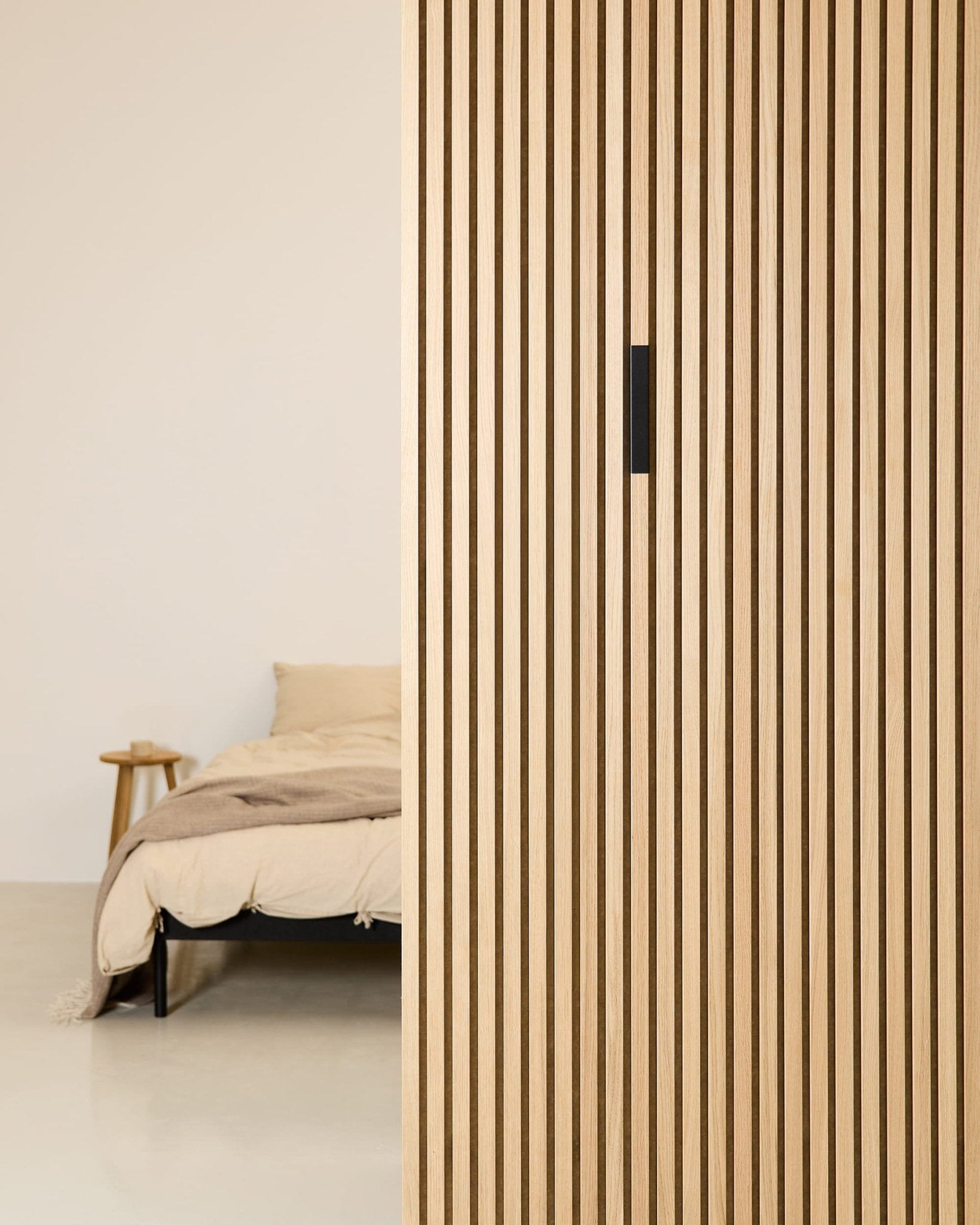 Et minimalistisk soveværelse med en beige seng, et sidebord i træ og et WoodUpp Liva Black Aluminium-akustikpanel, der tilfører tekstur til den lodrette skillevæg.