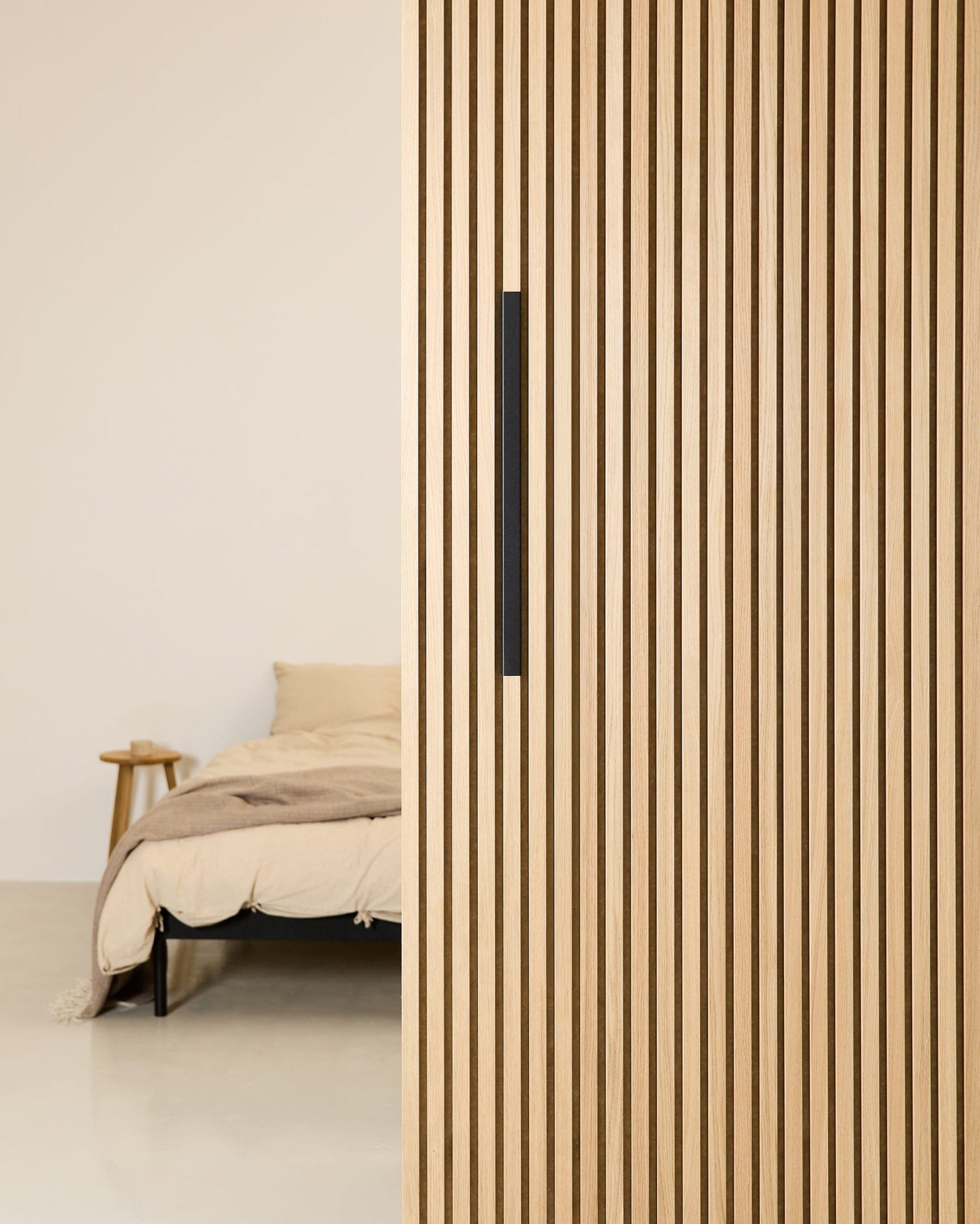 Minimalistisk soveværelse med en beige seng, der delvist afskærmes af en 60 cm bred WoodUpp EMBRACE rumdeler i sort aluminium med et sort håndtag.