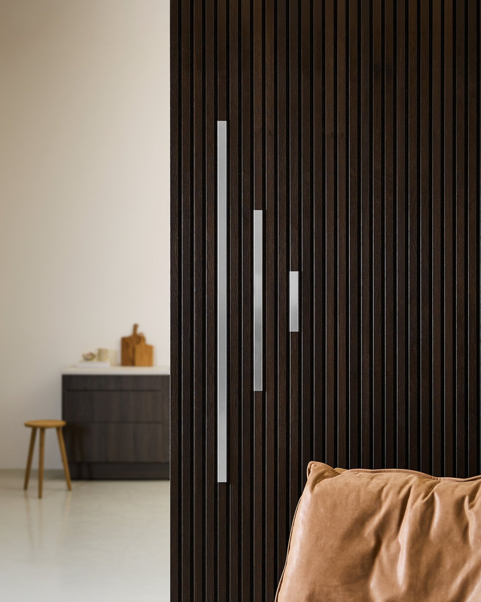 Minimalist interior featuring a dark wood Akupanel acoustic panel wall, WoodUpp Liva cover in polished steel (20 cm), tan cushion, and modern furniture.