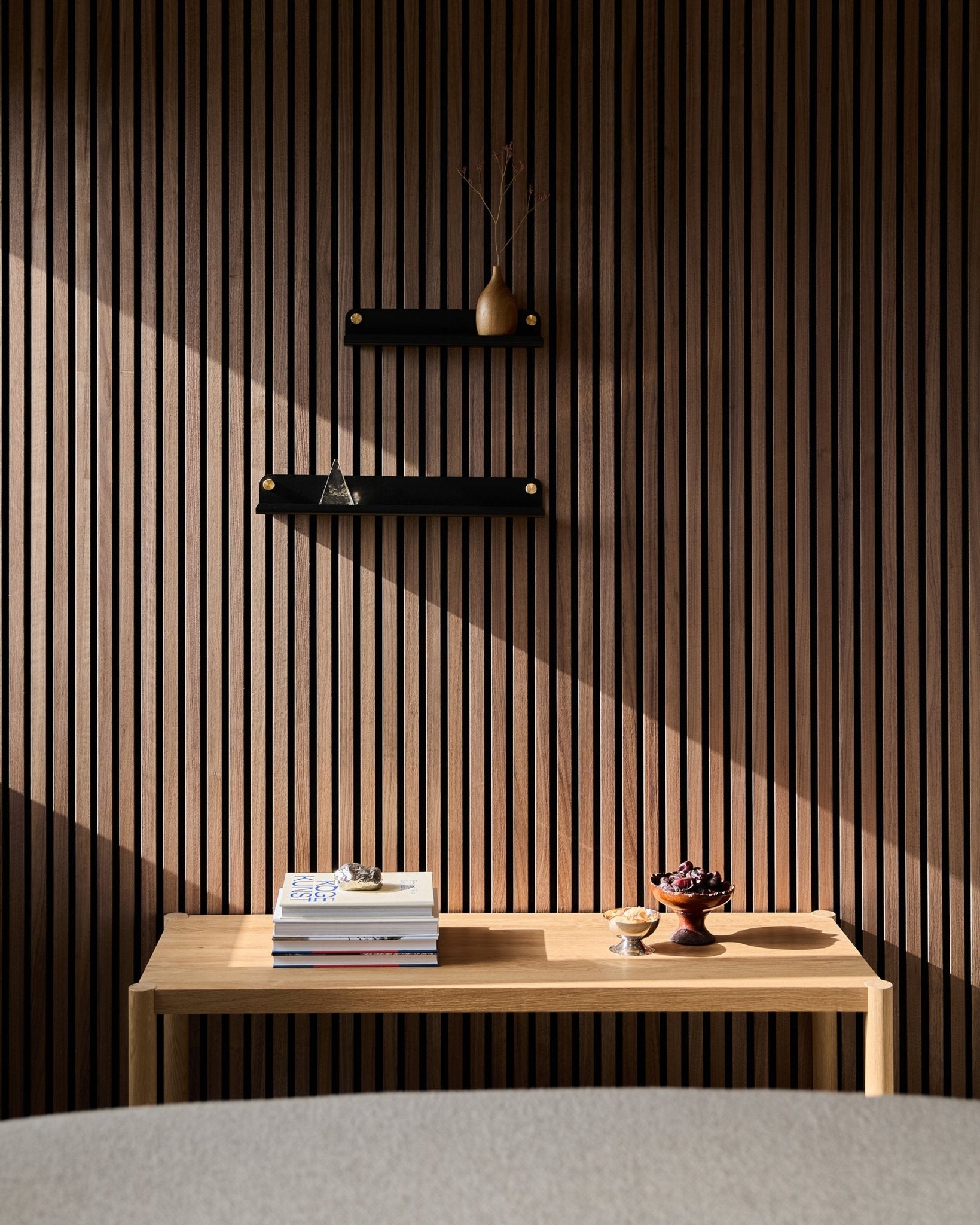 Et minimalistisk træbord med bøger og skåle står ved et nordisk akustikpanel af lameller, som fremhæver WoodUpp Norr Narrow Shelf i sort (51,9 cm); solbeskinnede skygger ovenover.