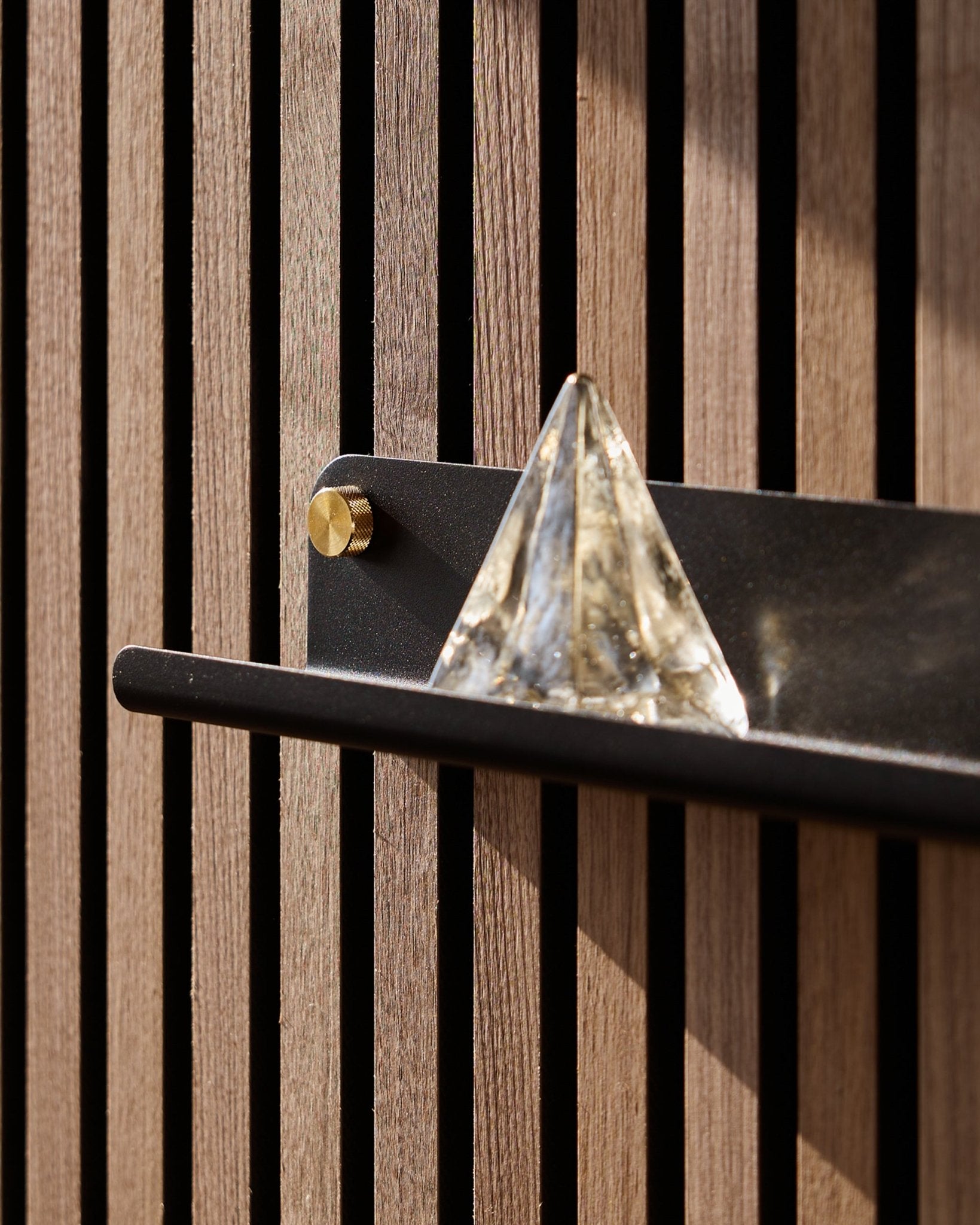 En glaspyramide står på WoodUpp Norr Narrow Shelf i black (51,9 cm), monteret på en vertical Akustikpanel og fremhævet af sollys.