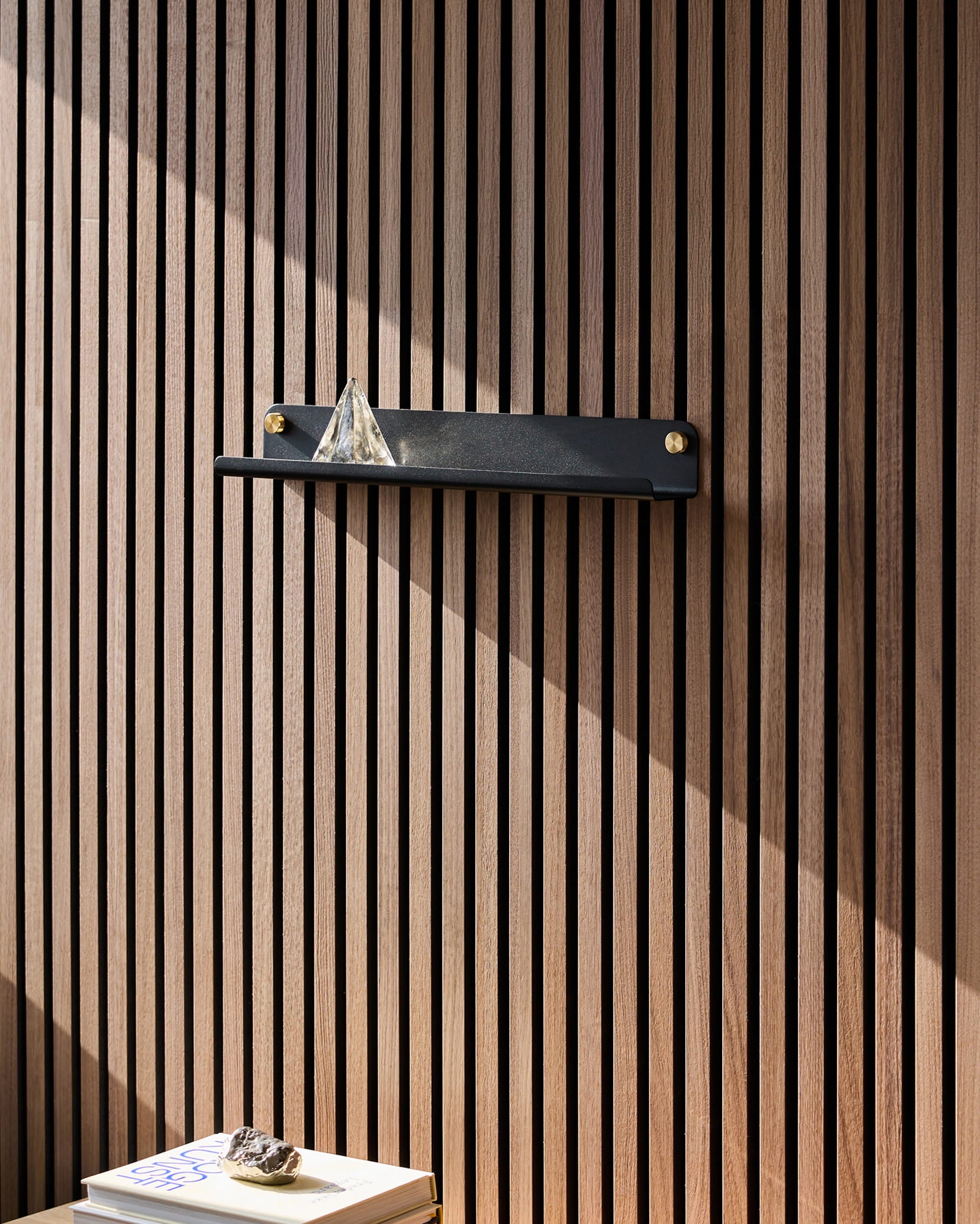 En sort Norr Narrow Shelf fra WoodUpp, monteret på et Akupanel-akustiktræpanel, bærer en glaspyramide; sollyset falder på bøgerne nedenunder, hvorpå der ligger en sten.