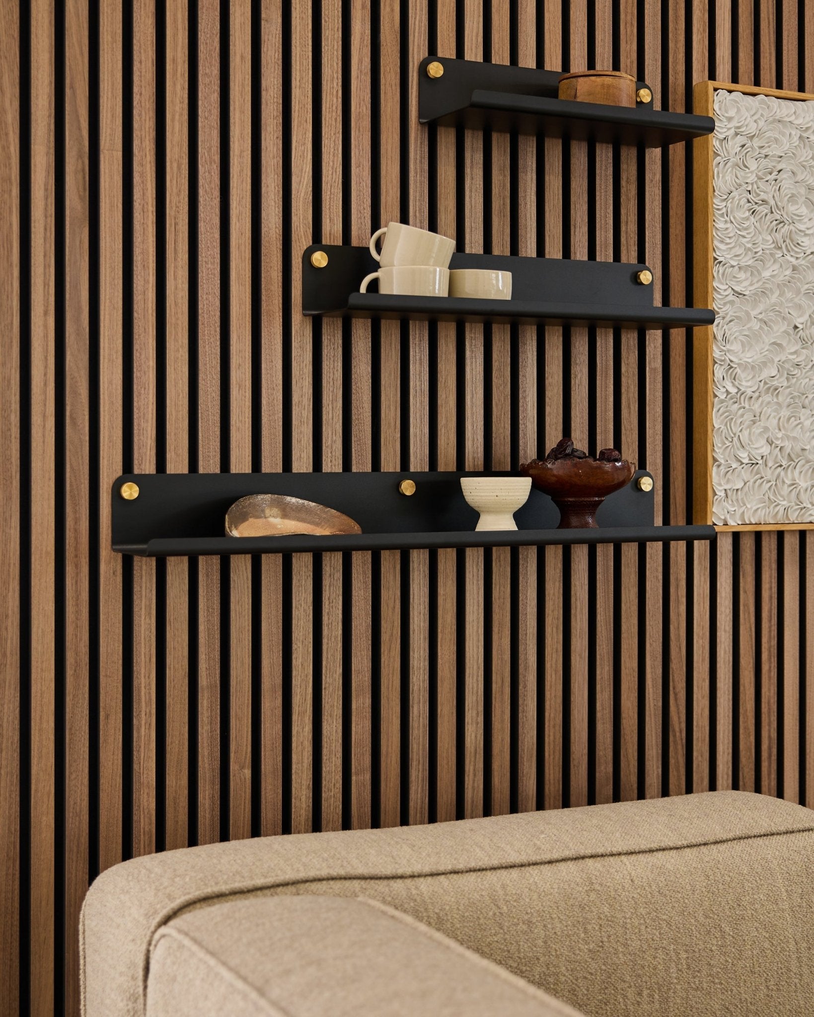 Tre sorte hylder, inklusive WoodUpp Norr Wide Shelf (75,9 cm), viser indretning på en akustisk panelvæg af trælameller over en beige sofa; en del af kunstværket er synligt.