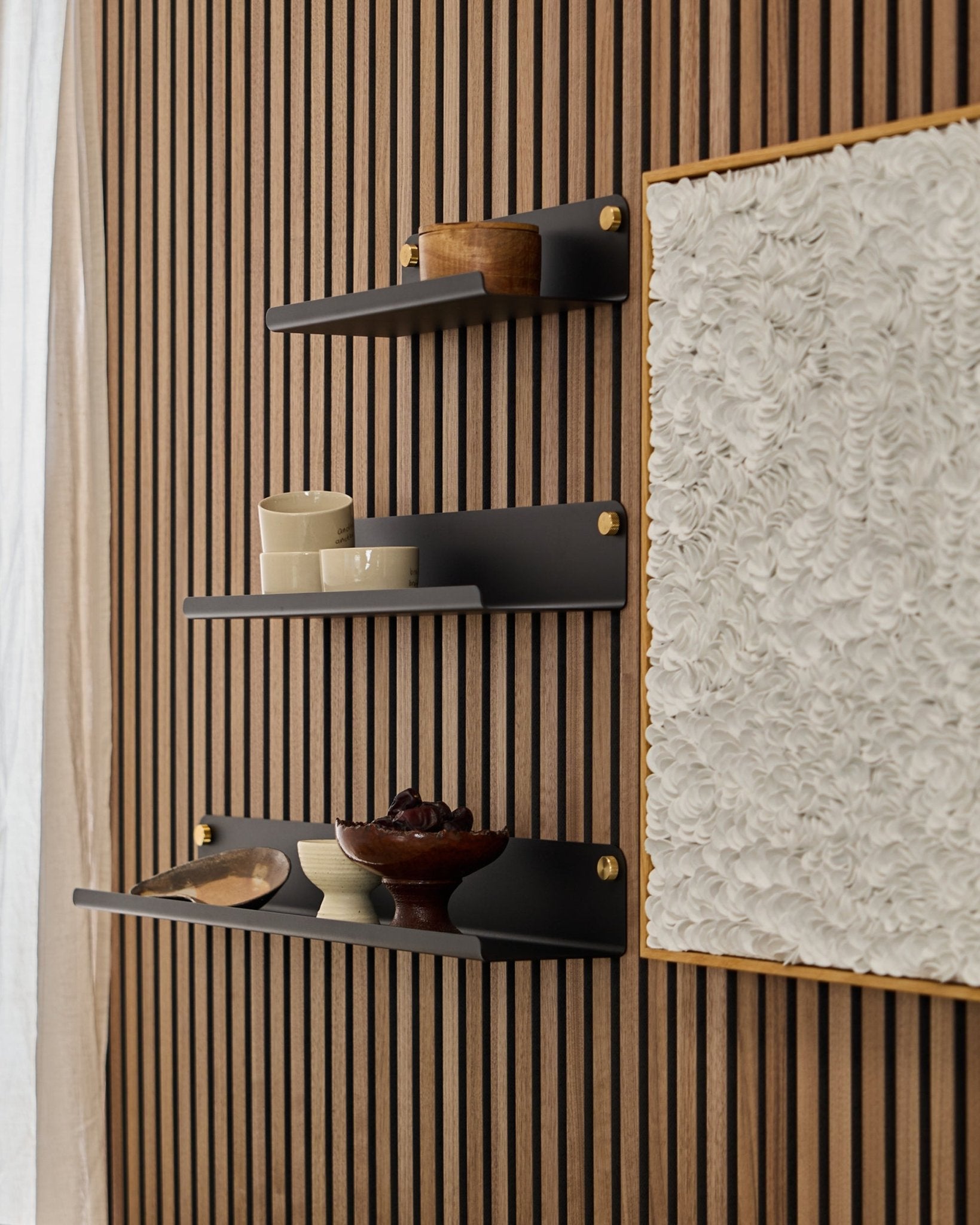 Three black WoodUpp Norr Wide Shelves (51.9 cm) mounted on a WoodUpp Akupanel acoustic wood panel, beside textured white art, displaying bowls and wood containers.