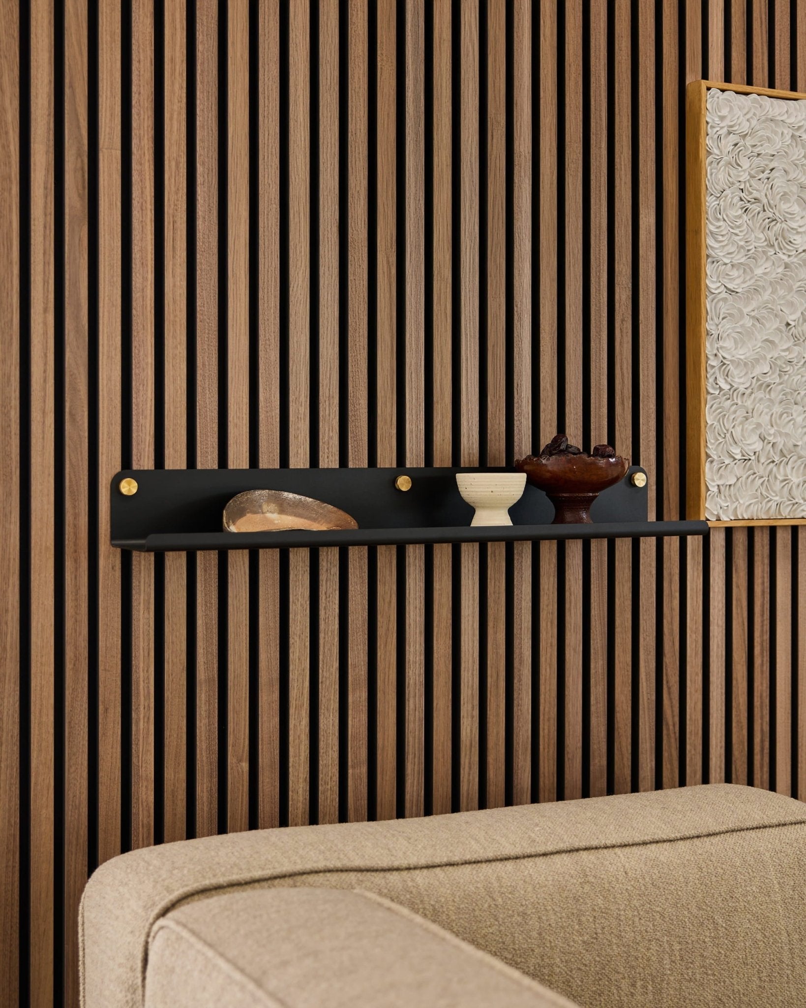 Sort WoodUpp Norr bred hylde (75,9 cm) med dekoration over en beige sofa, monteret på et moderne akustisk vægpanel af træ med lameller.
