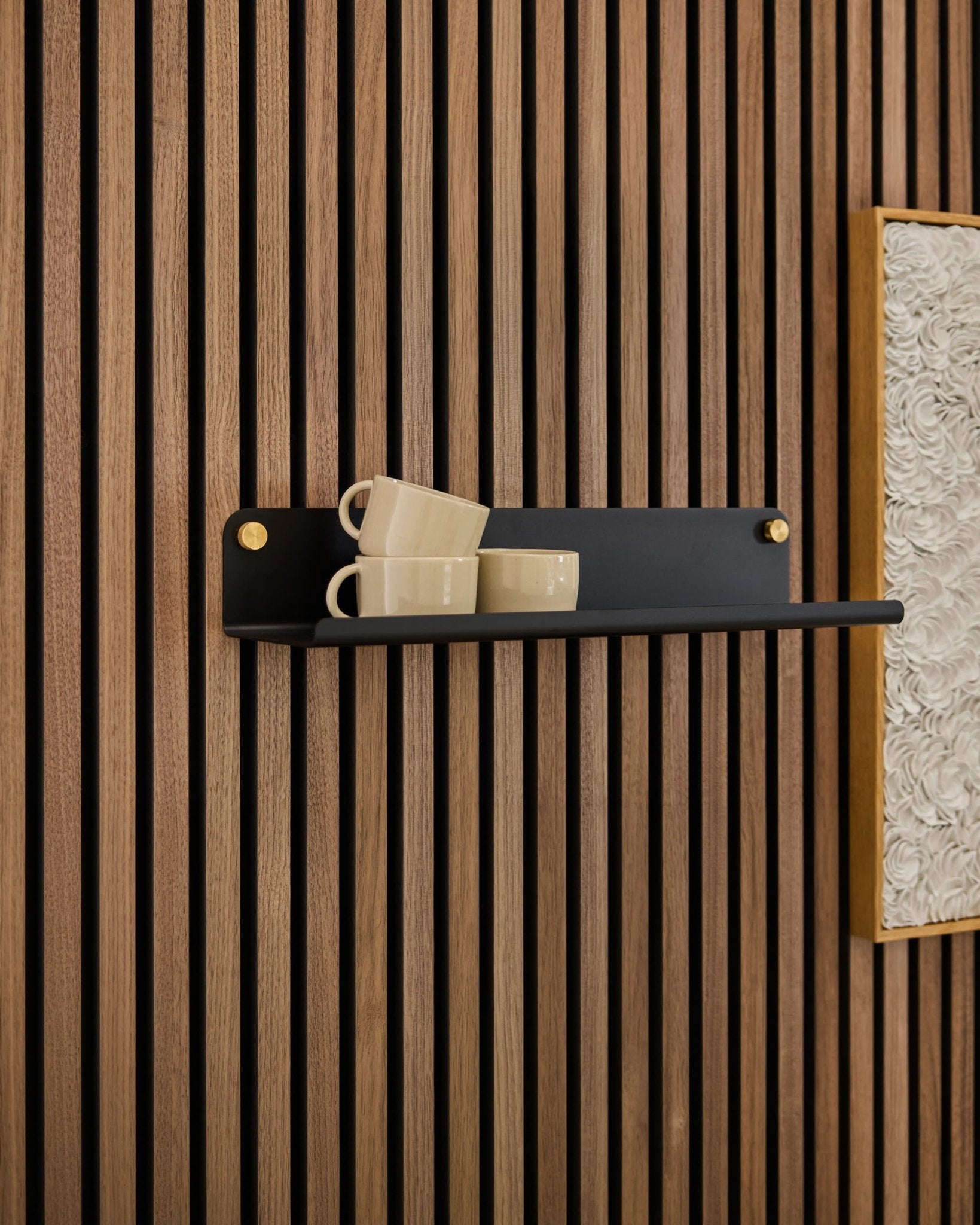 En sort Norr Wide Shelf fra WoodUpp rummer tre beige krus, der er monteret på et Akupanel-akustikpanel ved siden af et indrammet, hvidt kunstværk med struktur.