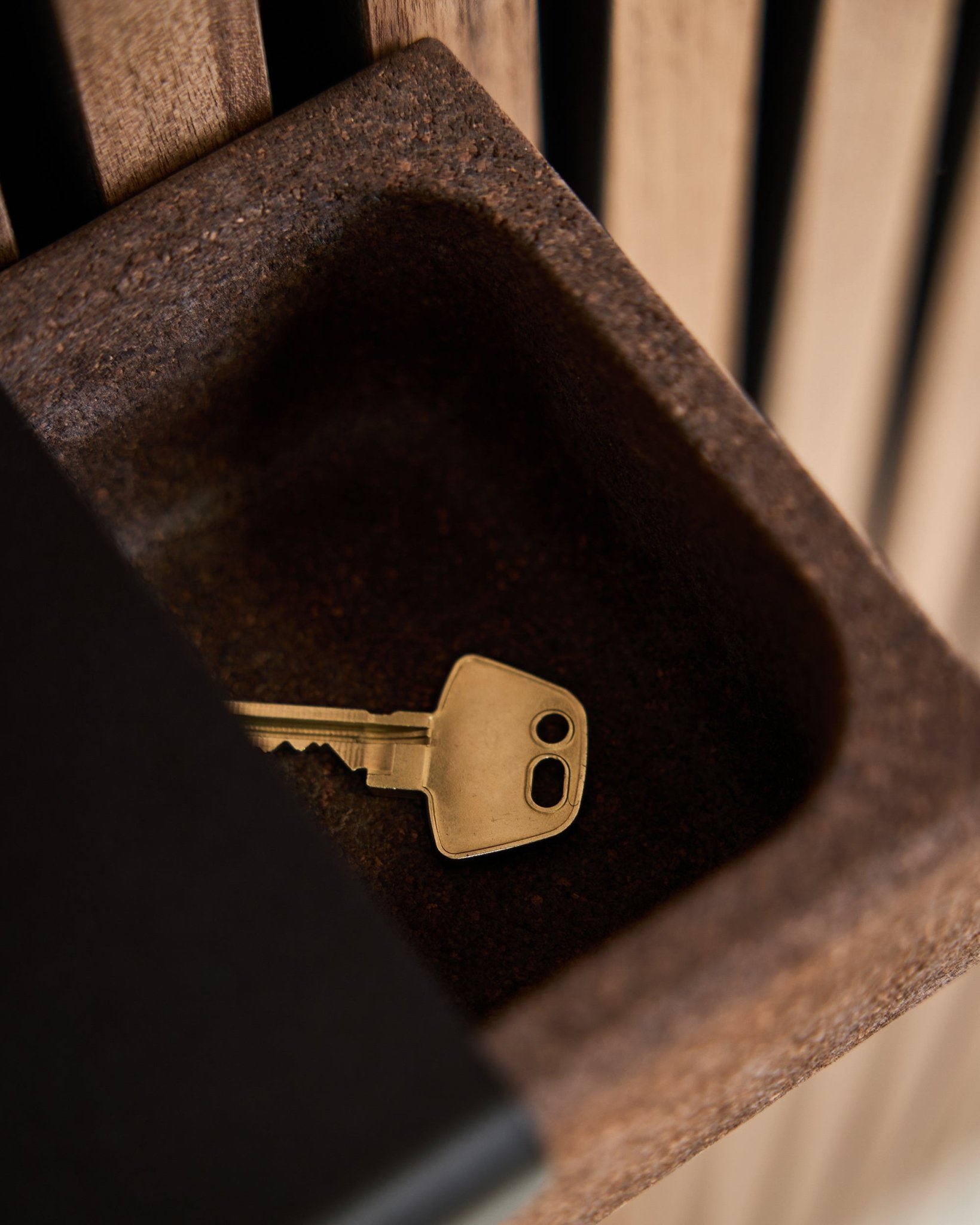 En brass nøgle ligger i en Norr Key Tray/røget kork, 35,9 cm fra WoodUpp, placeret på en brown beholder foran en baggrund af Akupanel akustiktræpanel.