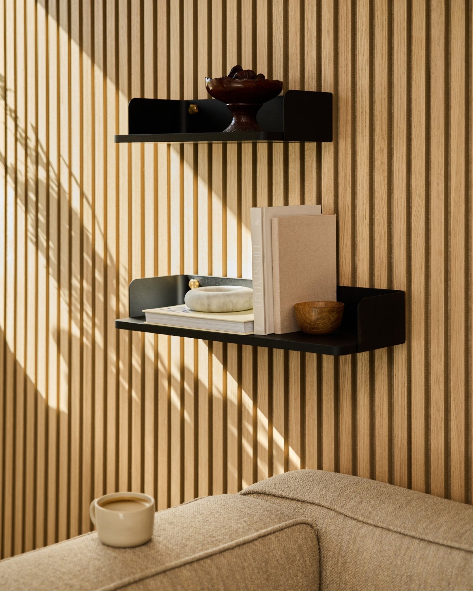 En black oak WoodUpp-boghylde (55,9 cm) viser bøger, dekoration og en skål på en væg med akustikpanel i træ; en sofa i beige og et krus ses i forgrunden.