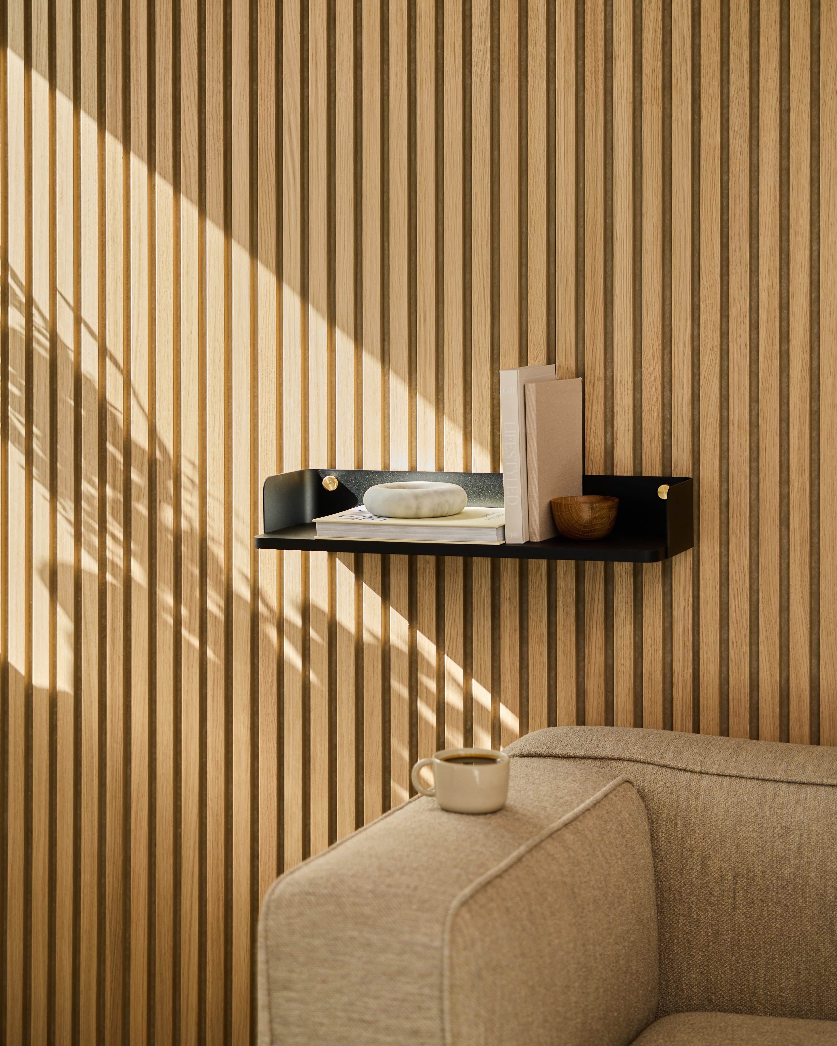 En Linn | Bogreol / Black Oak fra WoodUpp pryder væggen over en beige sofa. Sollyset kaster skygger på den stribede væg beklædt med akustiske træpaneler.