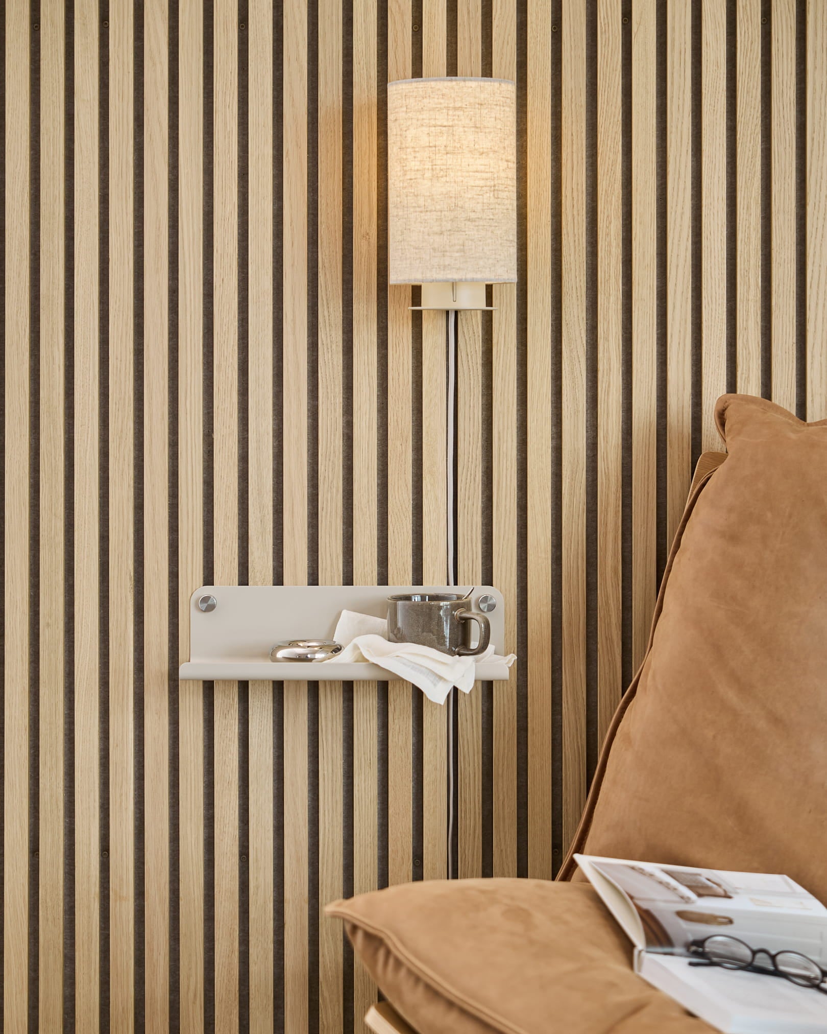 Lodret Akupanel-væg med akustiske trælameller, beige væglampe, lille hylde med kop og serviet, lysebrun sofa med et åbent magasin.