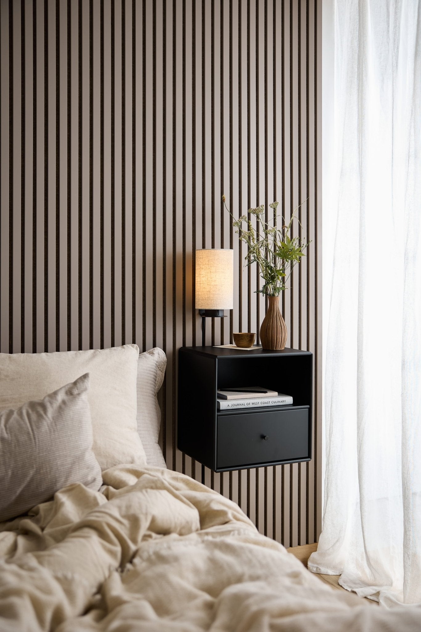 Seng med beige sengetøj, sort WoodUpp Arka bogreol med skuffe, lampe og vase ved et vindue med tynde hvide gardiner.