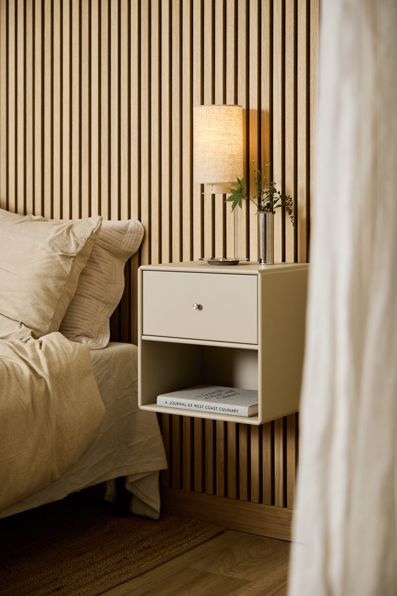 En beige WoodUpp Arka-reol med skuffe ved sengen rummer en lampe, bøger og en vase og står foran et WoodUpp Akupanel-akustisk trælamelpanel.