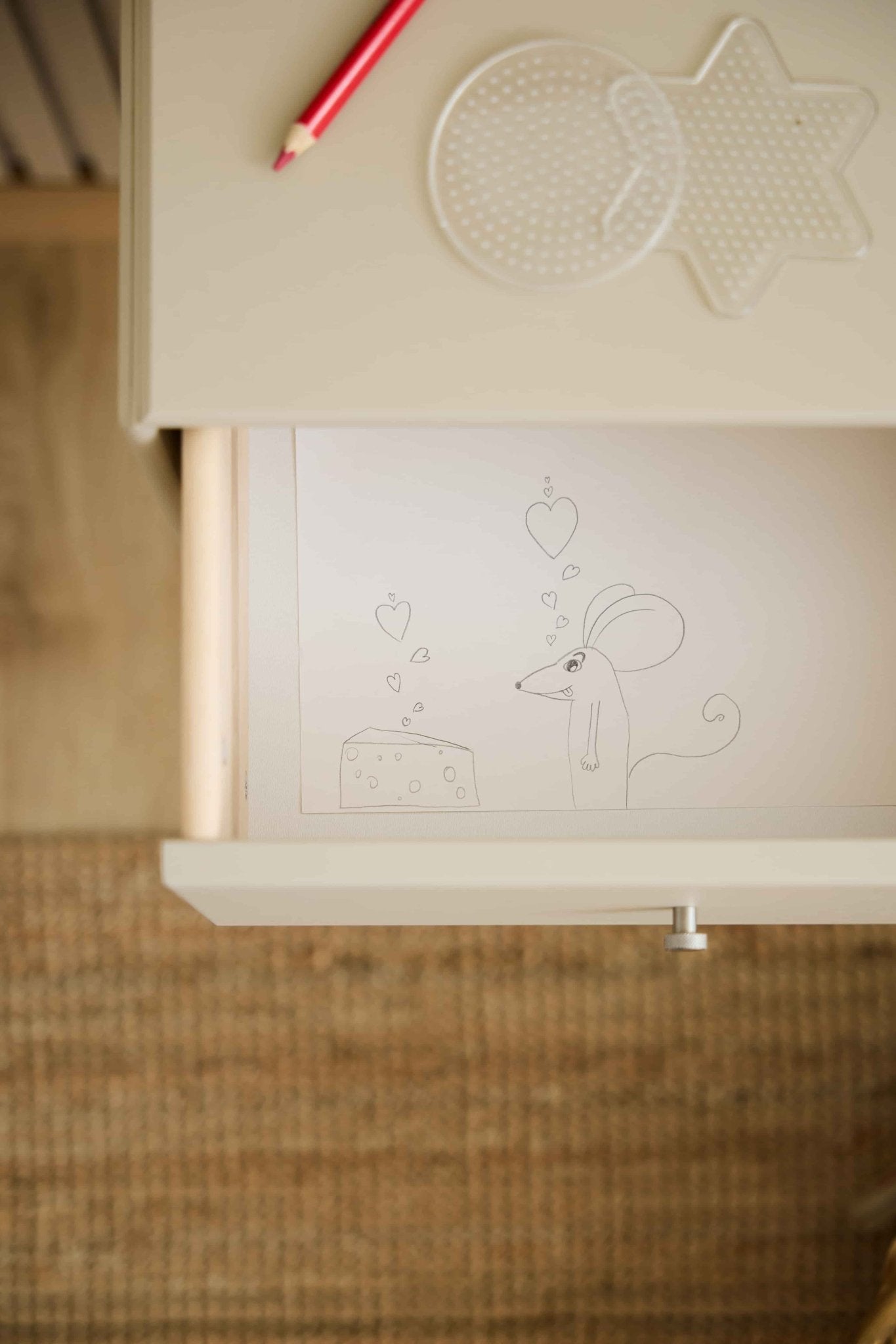 En mus og et stykke ost med hjerte-motiv pryder en WoodUpp Arka-reolskuffe i beige; en rød blyant og nogle hulplader ligger på den matchende bordplade ovenover.
