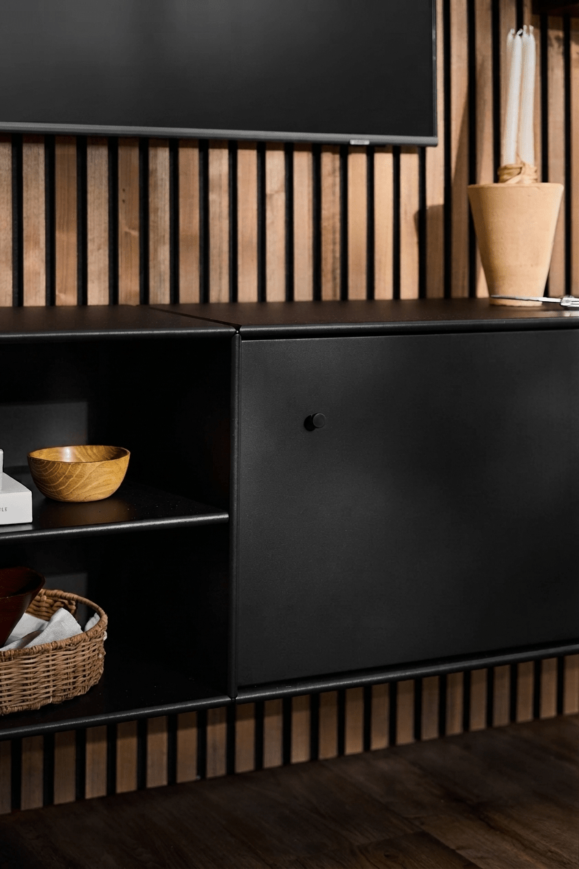 Sort WoodUpp Arka TV-kommode med åbne hylder til bøger og kurve, en beige vase, smart kabelstyring og en lodret Akupanel-akustikvæg af træ som baggrund.