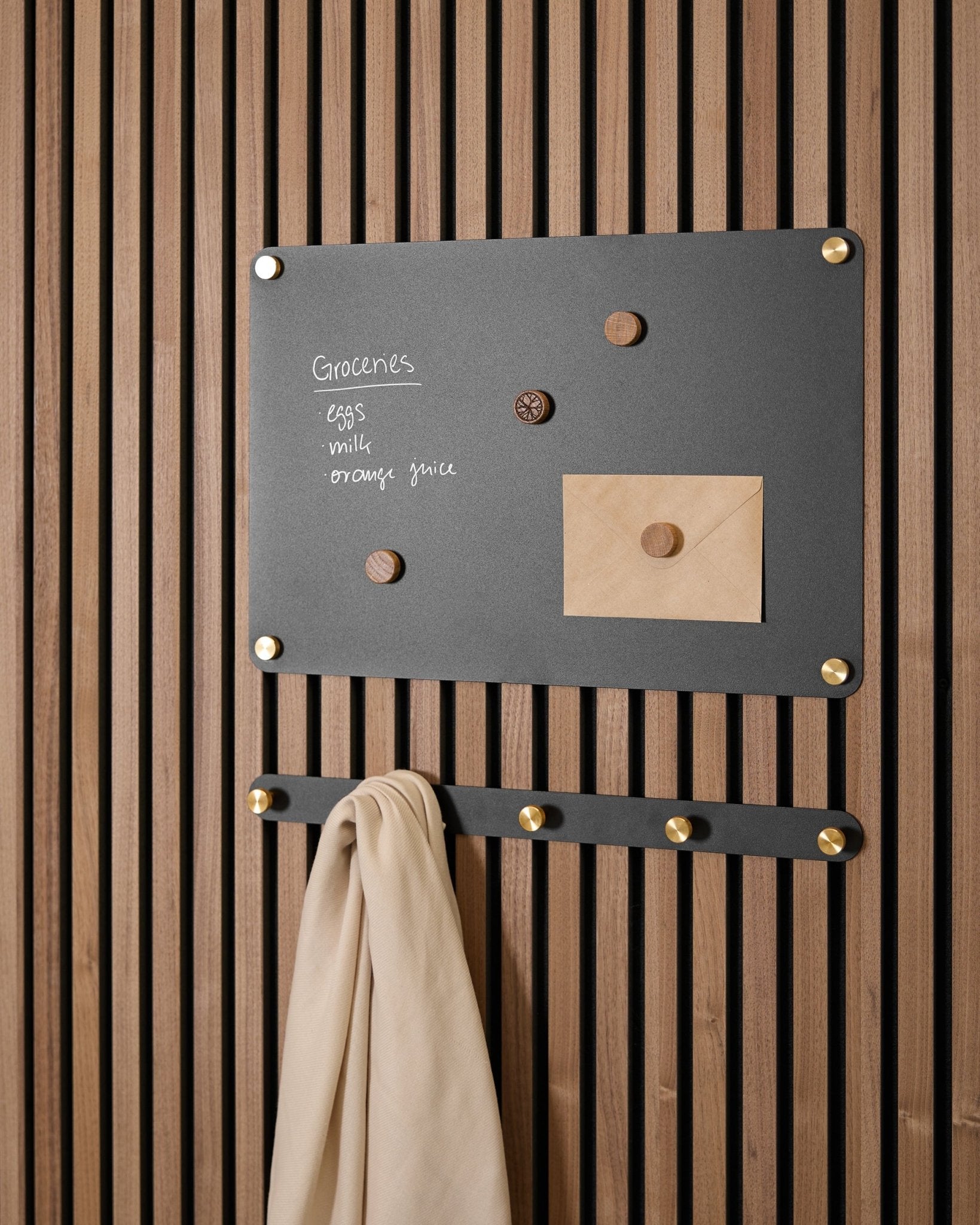 En small black Norr magnettavle fra WoodUpp holder en indkøbsliste, en kuvert, magneter og beige stof på kroge og er monteret på en Akupanel Akustikpanel.