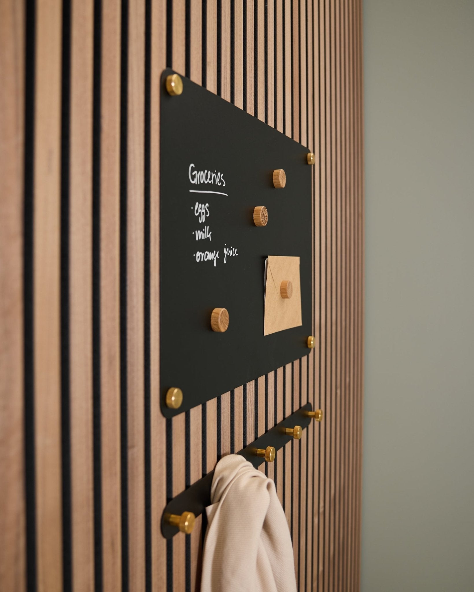 En small black Norr magnettavle fra WoodUpp monteret på et Akupanel akustisk træpanel viser en indkøbsliste og en note, med kroge nedenunder, der holder et lyst stykke stof.