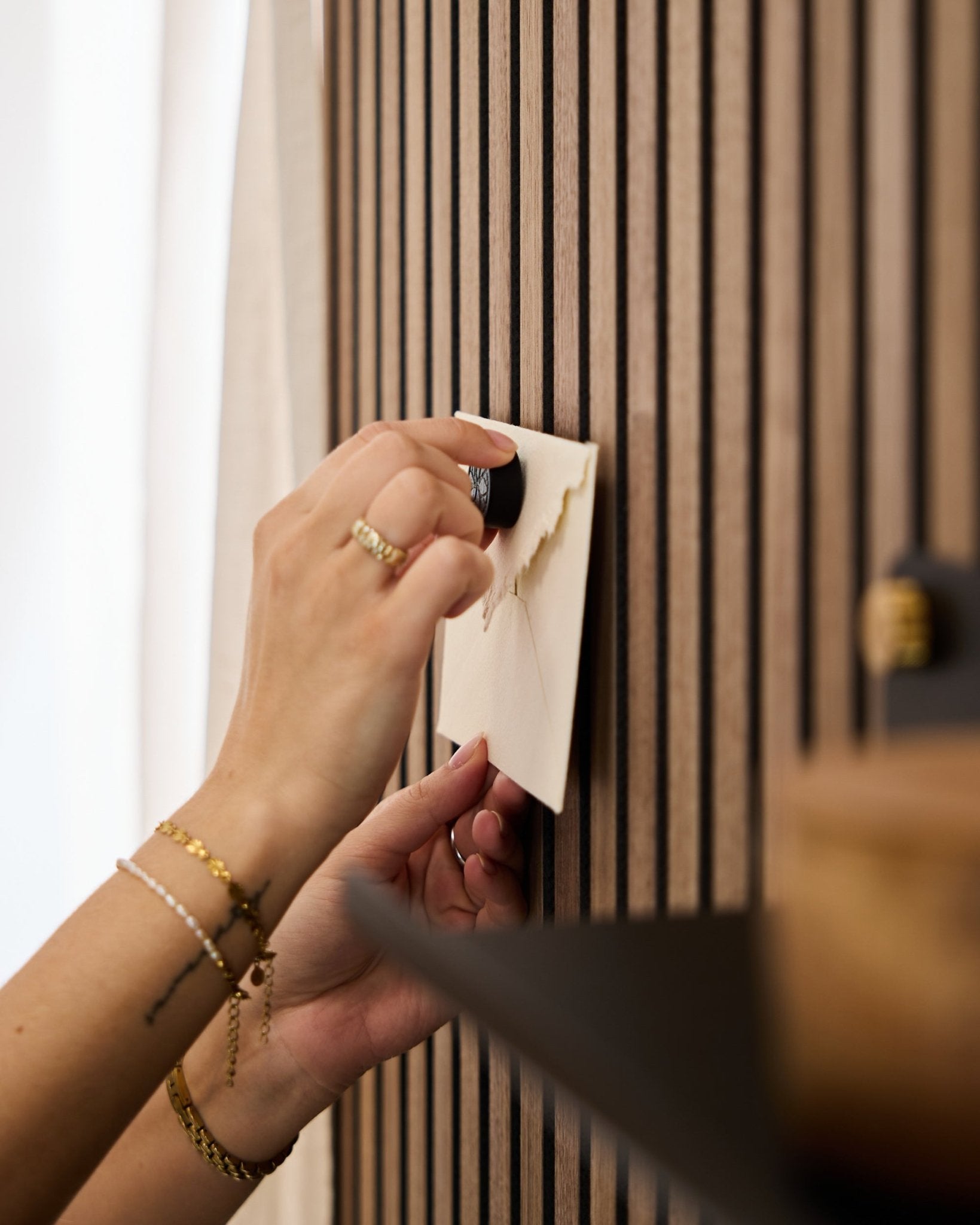 En person med guldbånd og ringe sætter en kuvert fast på et WoodUpp akustikpanel med egetræslameller ved hjælp af Norr Poster Magnet-sættet i Black Oak.