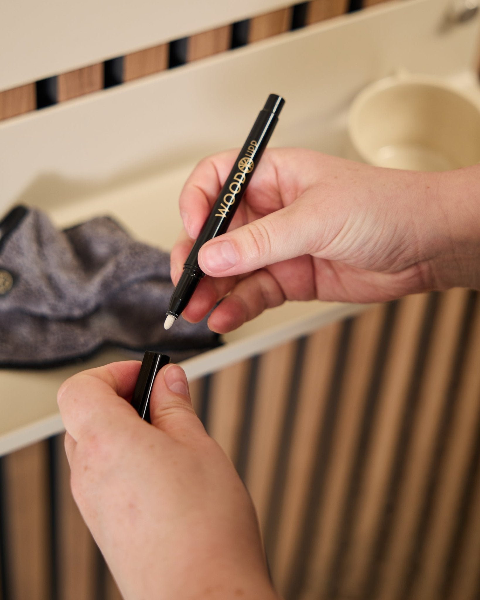 En person holder en åben WoodUpp Norr | Blackboard set tusch ved siden af en mikrofiberklud og en beige kop, produkter designet til brug med akustiske træpaneler.