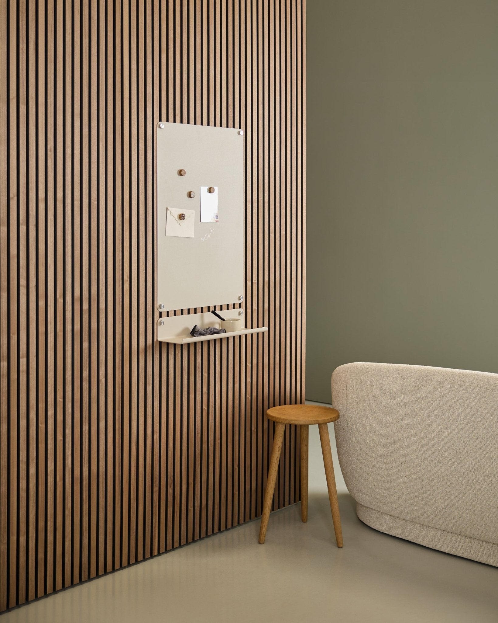 Minimalist interior with beige sofa and wooden stool, featuring a large beige WoodUpp Norr Magnetic Board mounted on a Nordic Akupanel acoustic wood panel near a green wall.