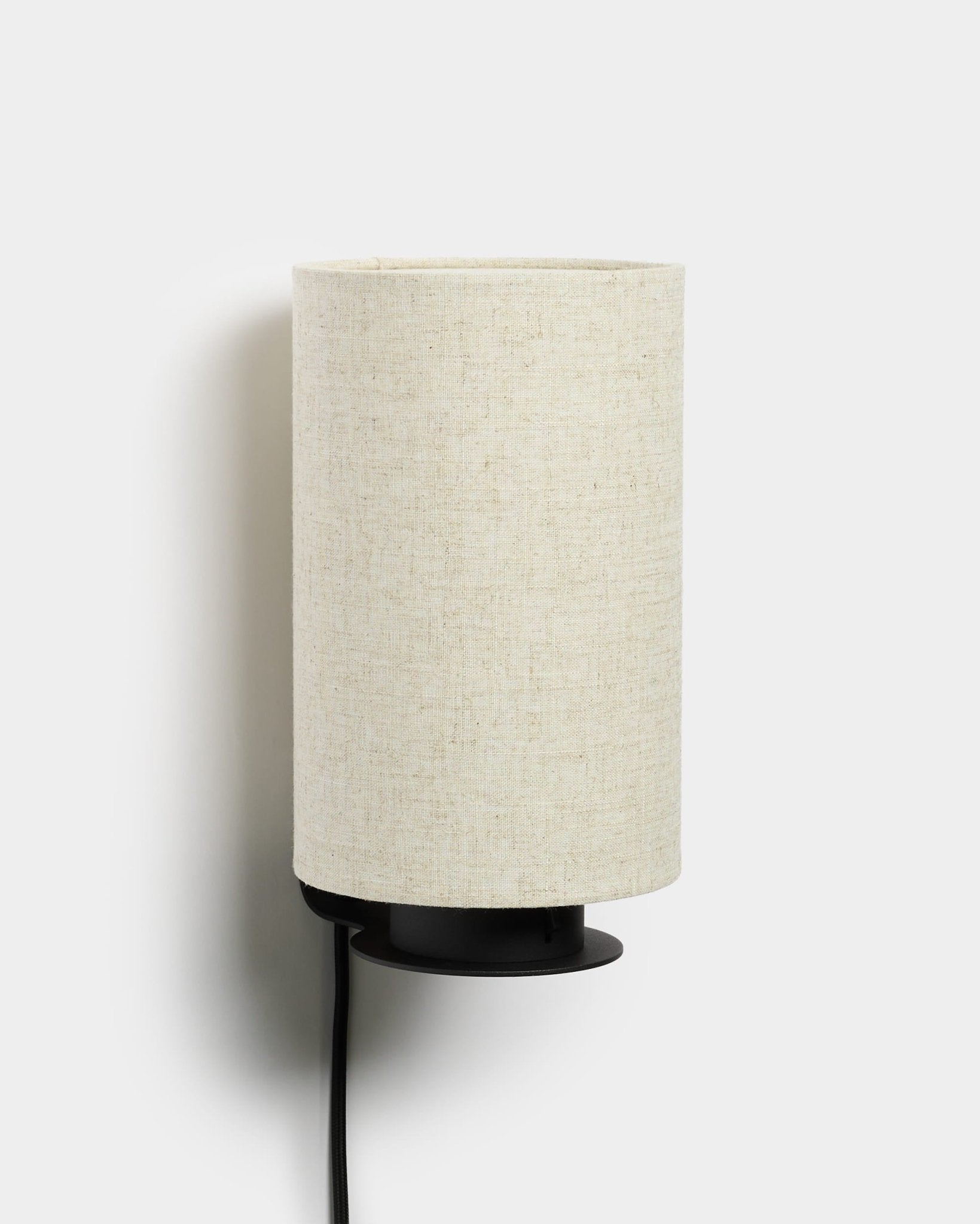 Feya væglampe fra WoodUpp med en sort metalbase, tekstureret hør-bomuldsskærm i beige og en synlig sort strømledning. Nordisk minimalistisk design.