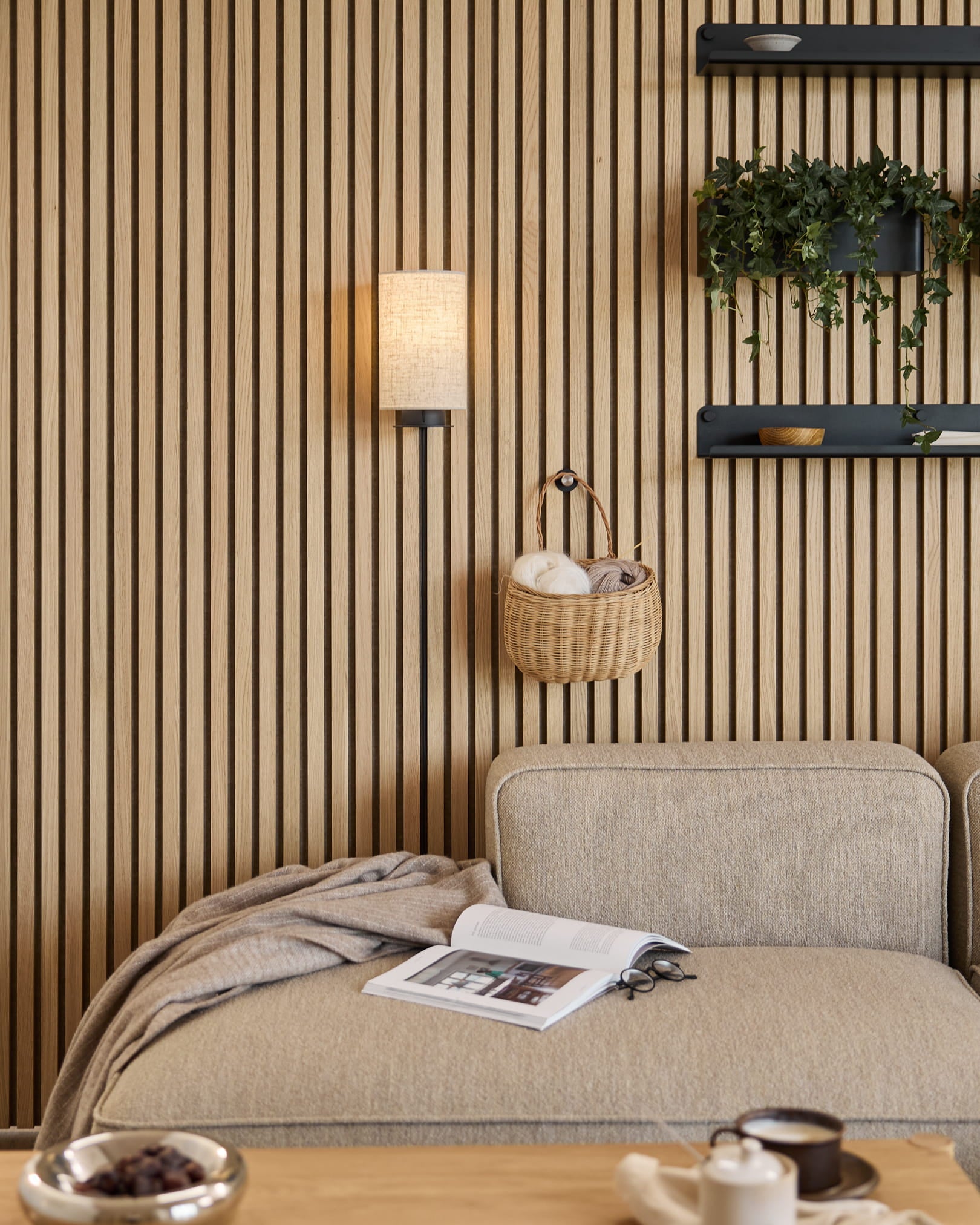 Beige sofa med tæppe og bog ved siden af en WoodUpp Akupanel akustisk lamelvæg, hylder med planter og dekorationer og en Feya-væglampe i sort/beige.