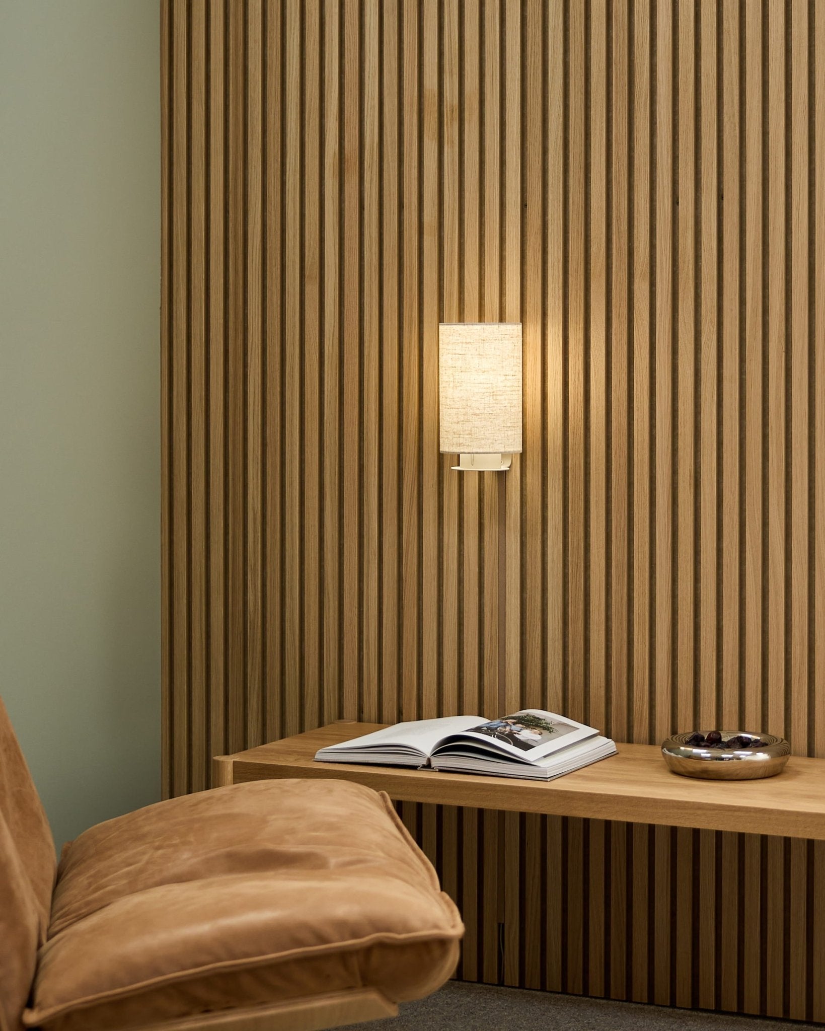 Minimalistisk nordisk rum med en tanfarvet stol, WoodUpp Akupanel akustisk lamelvæg, Feya væglampe i Beige/Beige, en åben bog på en bænk og en skål.