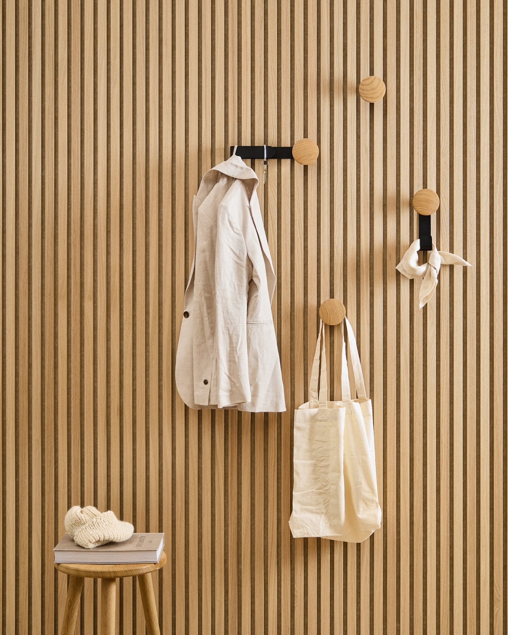 En beige jakke, taske og tørklæde hænger på WoodUpps Linn Horizontal Hanger i Natural Oak; en skammel ved siden af bærer en bog og en strikket genstand.