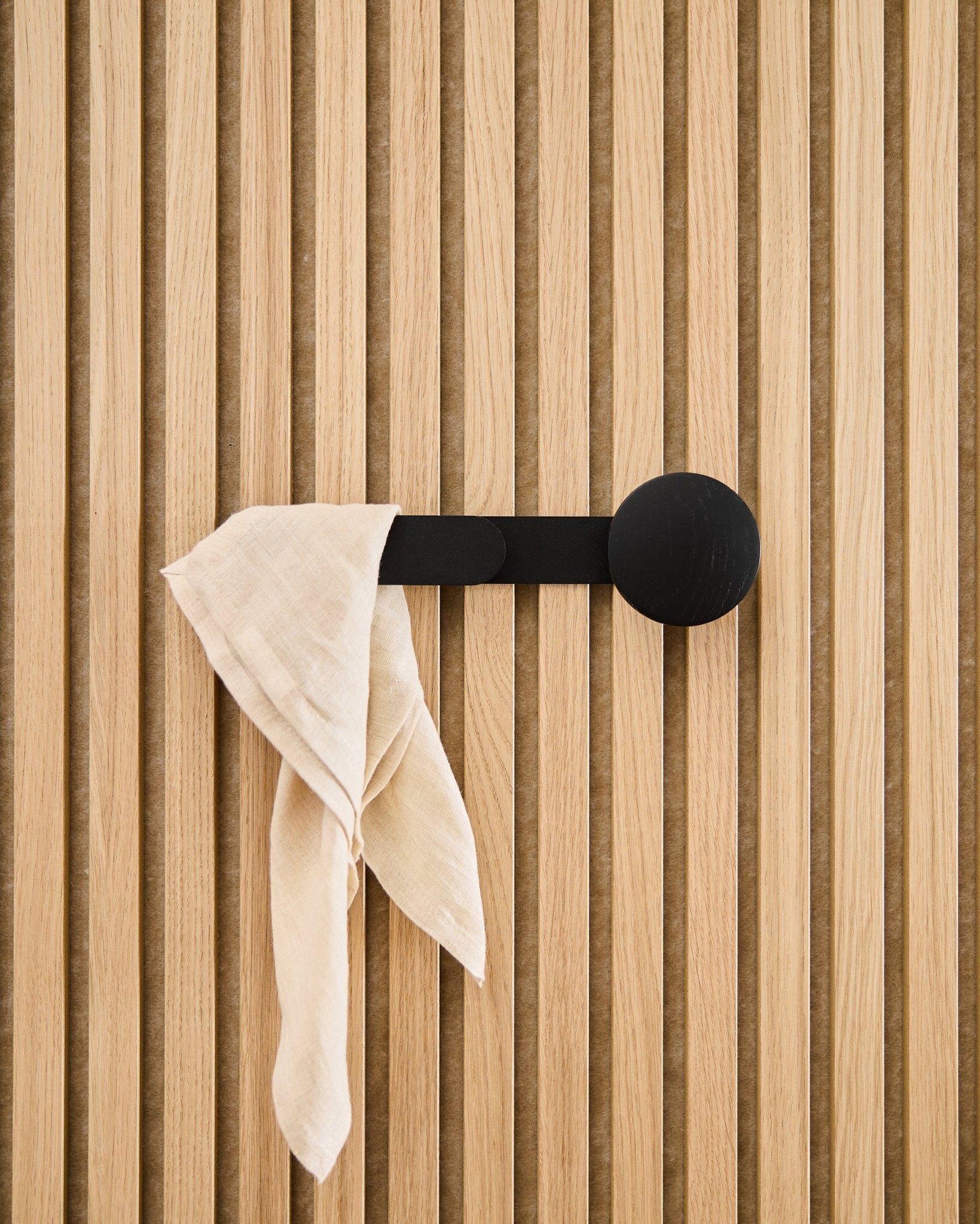 Et beige klæde hænger over WoodUpp Linn Horizontal Hanger i Black Oak, monteret på et vertical akustikpanel med trælameller.