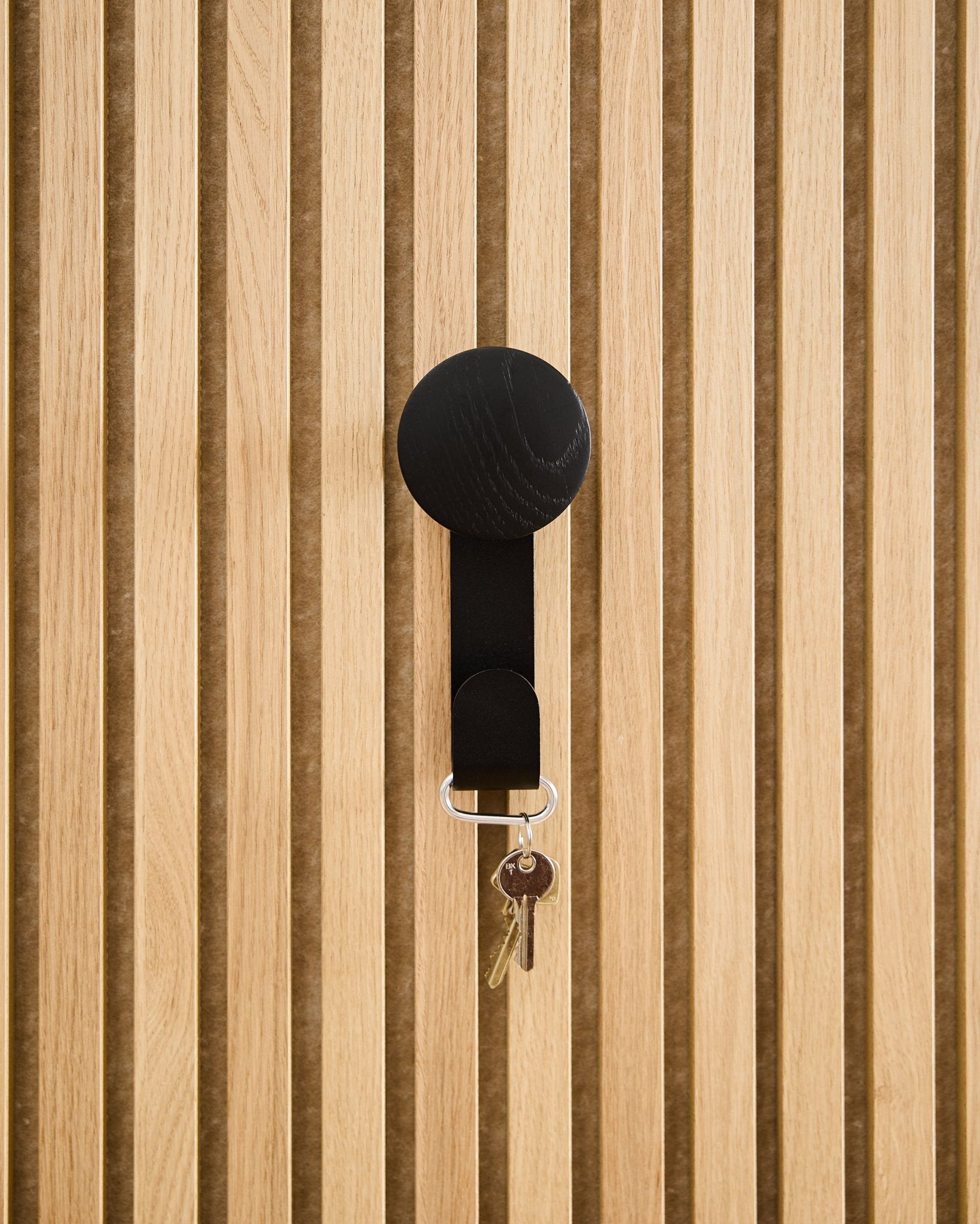Linn Vertical Hanger i sort eg fra WoodUpp holder en nøglering med to nøgler og et rundt mærke, monteret på et akustisk Akupanel-træpanel med struktur.