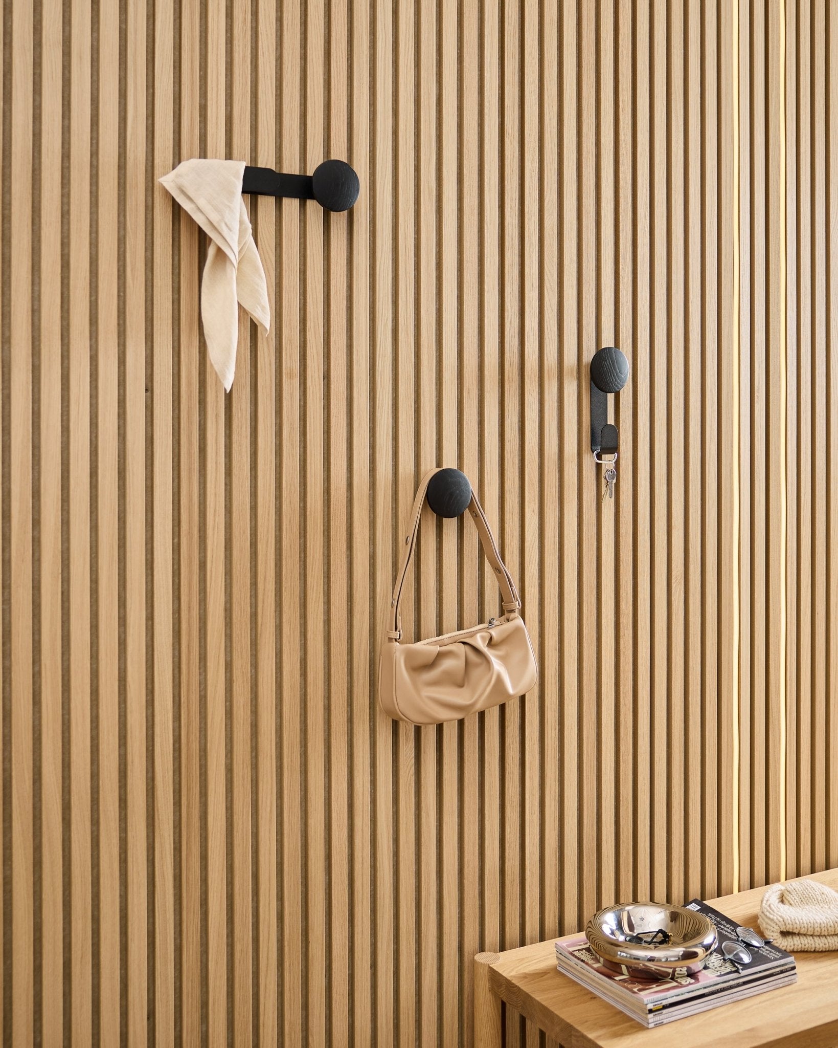 WoodUpp Linn Single Hanger i Black Oak holder et tørklæde, en beige taske og nøgler på et black Akupanel akustisk træpanel; bænk med bøger og metalskål i nærheden.