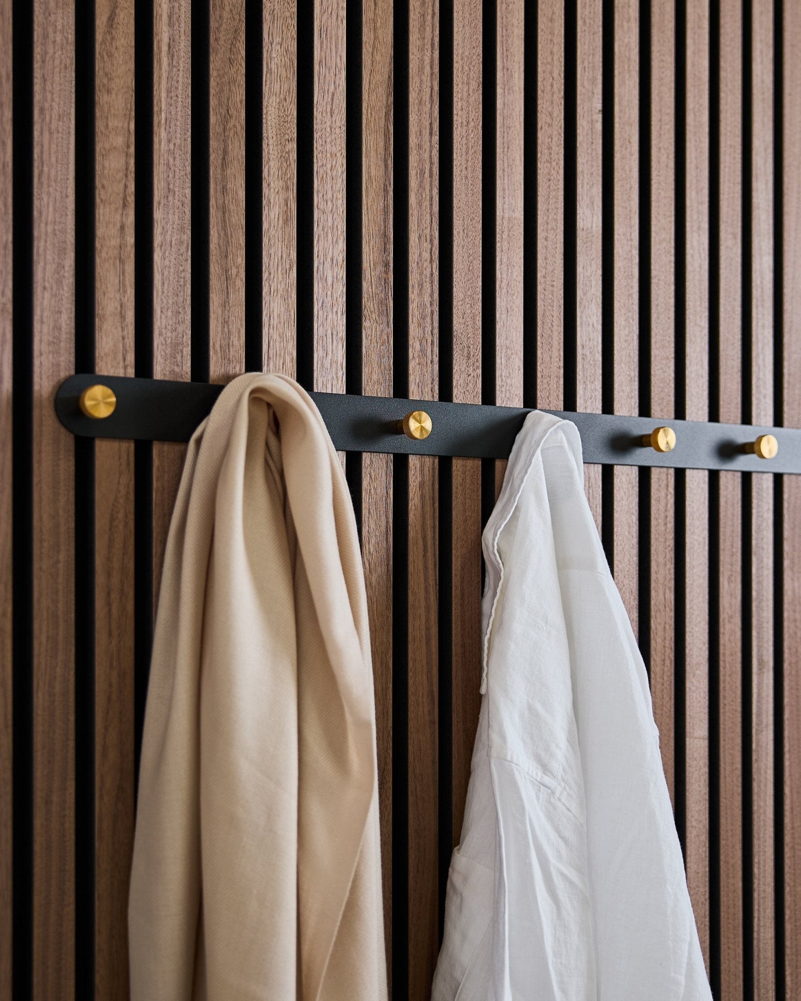WoodUpp Norr knagerække i black/brass (X7) med et beige tørklæde og en hvid skjorte hænger på et nordisk akustikpanel i trælameller.
