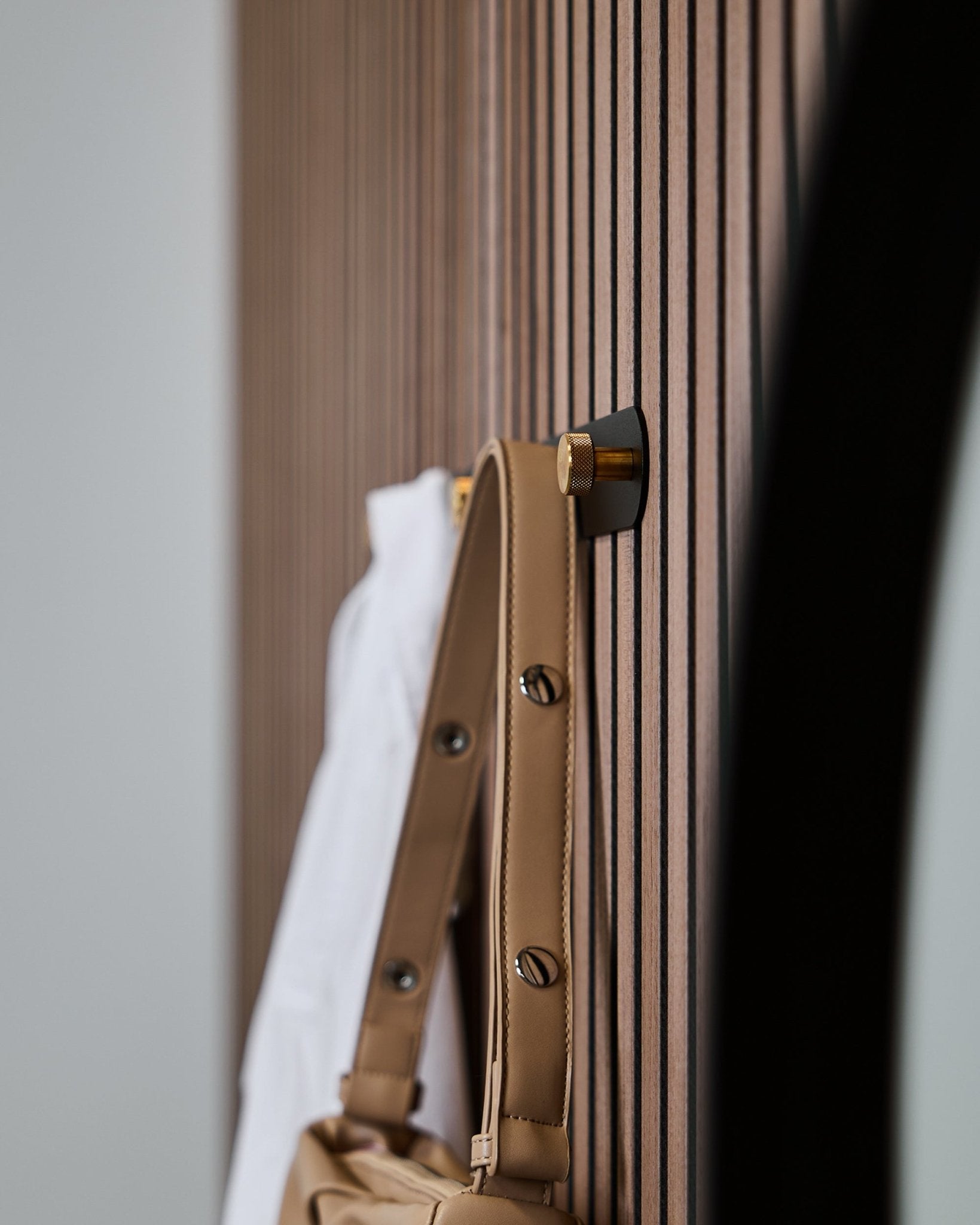 En lys brun taske og en hvid skjorte hænger på en WoodUpp Norr knagerække (Black/Brass, X5), monteret på et vertical Akupanel akustikpanel med trælameller.