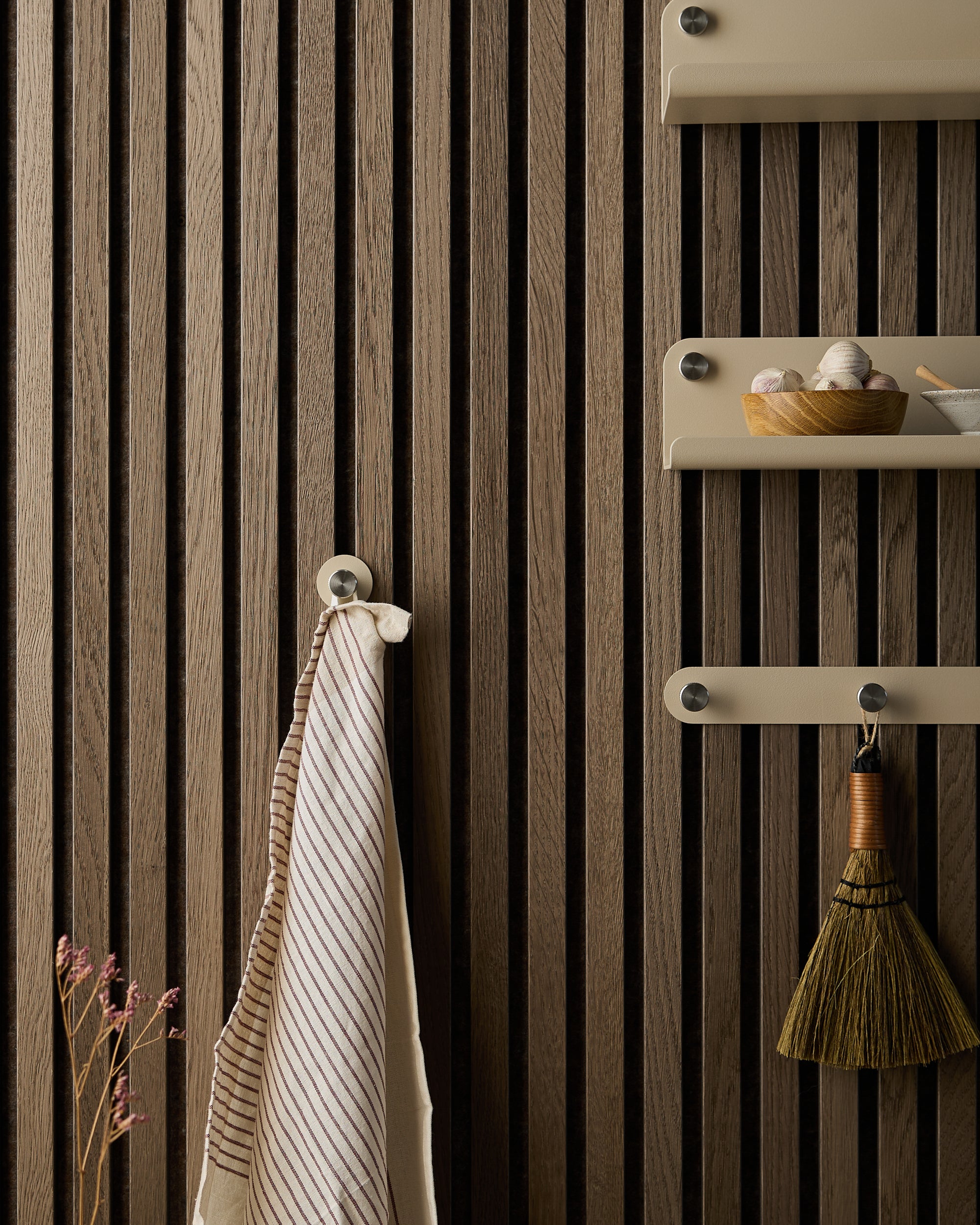 Striped towel, small broom, and bowl rest on shelves mounted to a dark Akupanel acoustic wood panel, with dried flowers at the bottom left.