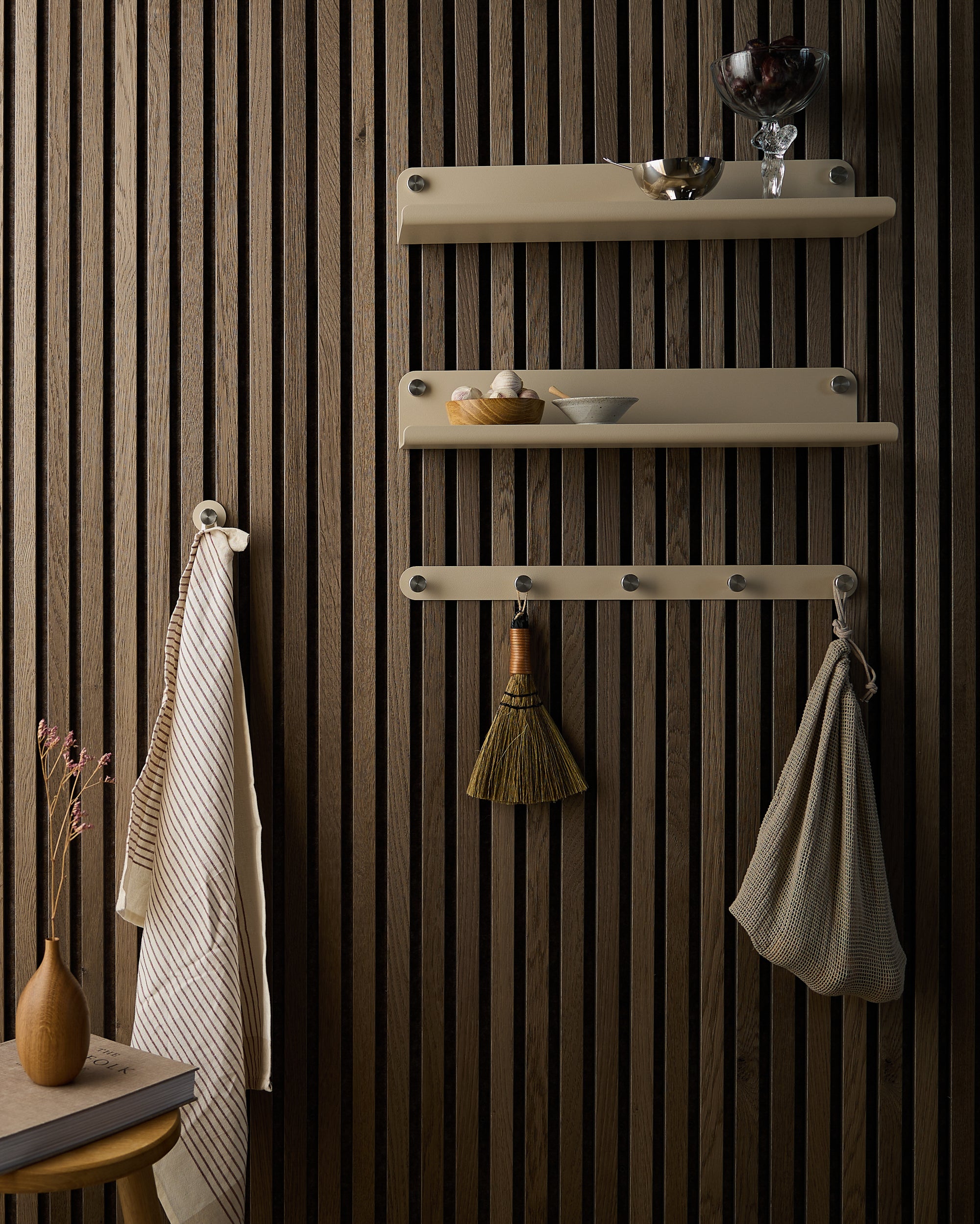 Vertical Nordic Akupanel acoustic wood slatwall with three beige dish shelves, hooks holding towels and broom; small table with vase nearby.