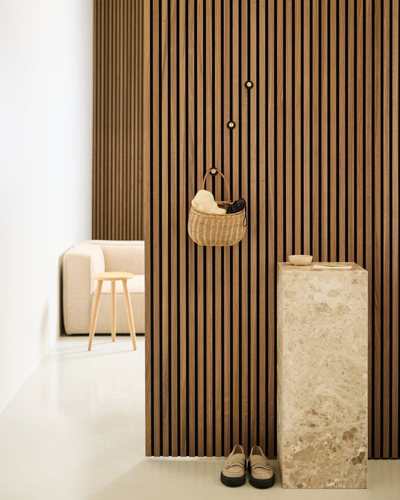 Minimalist entryway featuring Akupanel acoustic slat wall, WoodUpp Norr Coat Hook in black and brass, basket, stone pedestal, slippers, and beige chair.