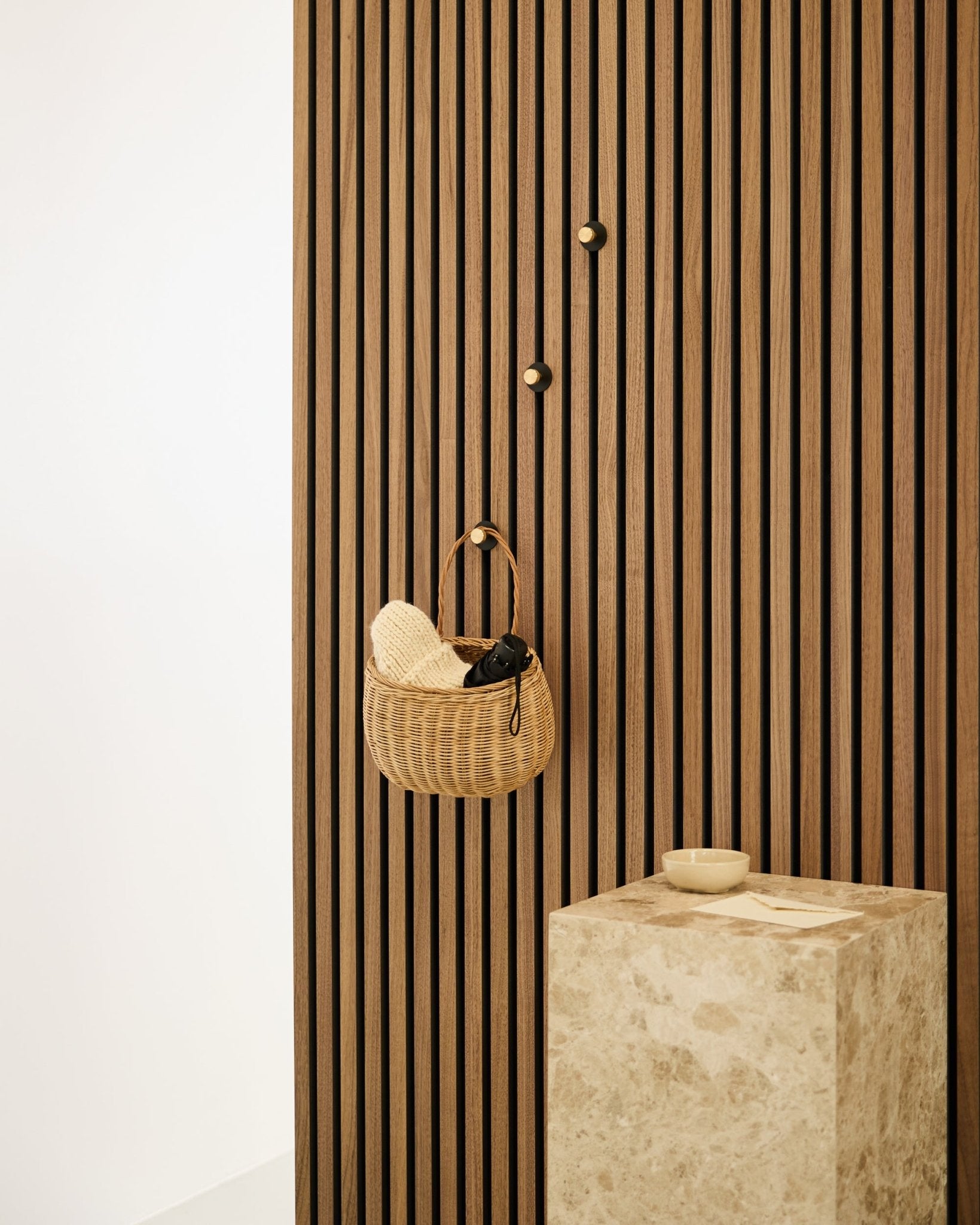 Norr Coat Hook fra WoodUpp monteret på en nordisk Akupanel akustisk lamelvæg ved siden af et beige stenpodie med en small skål i et minimalistisk interiør.