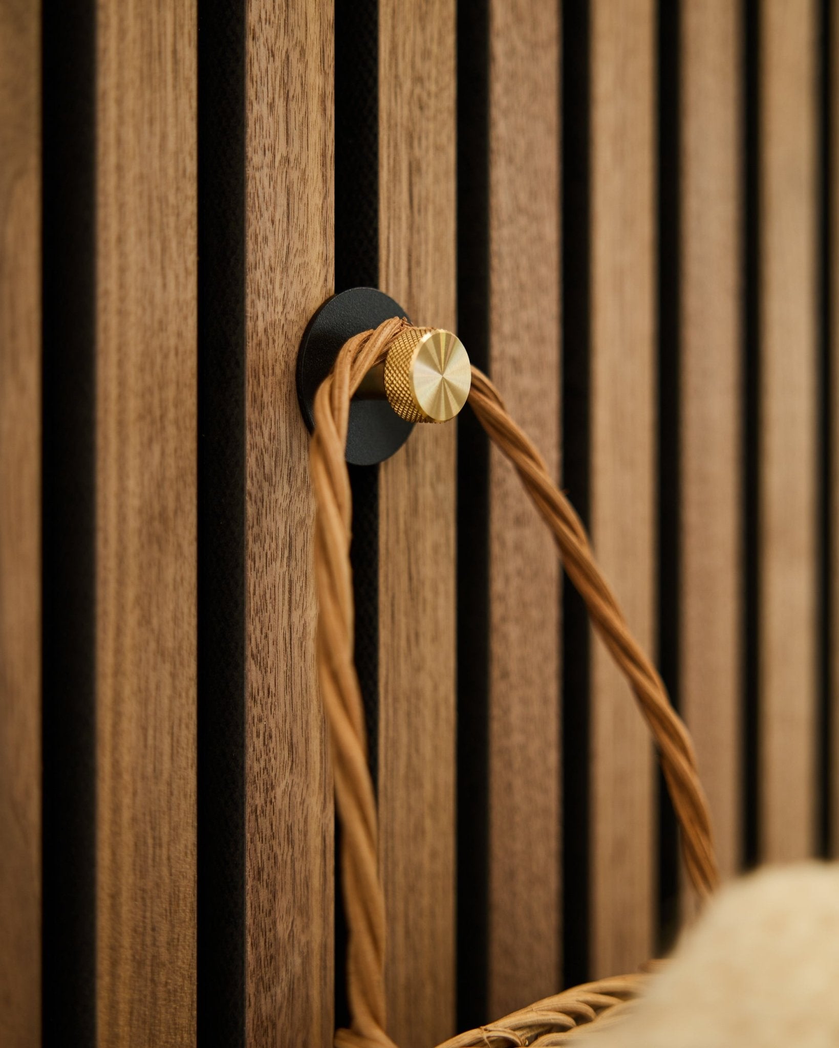 WoodUpp Norr knage i black og brass, monteret på en træbeklædt Akupanel Akustikpanel, holder et flettet håndtag i tan.