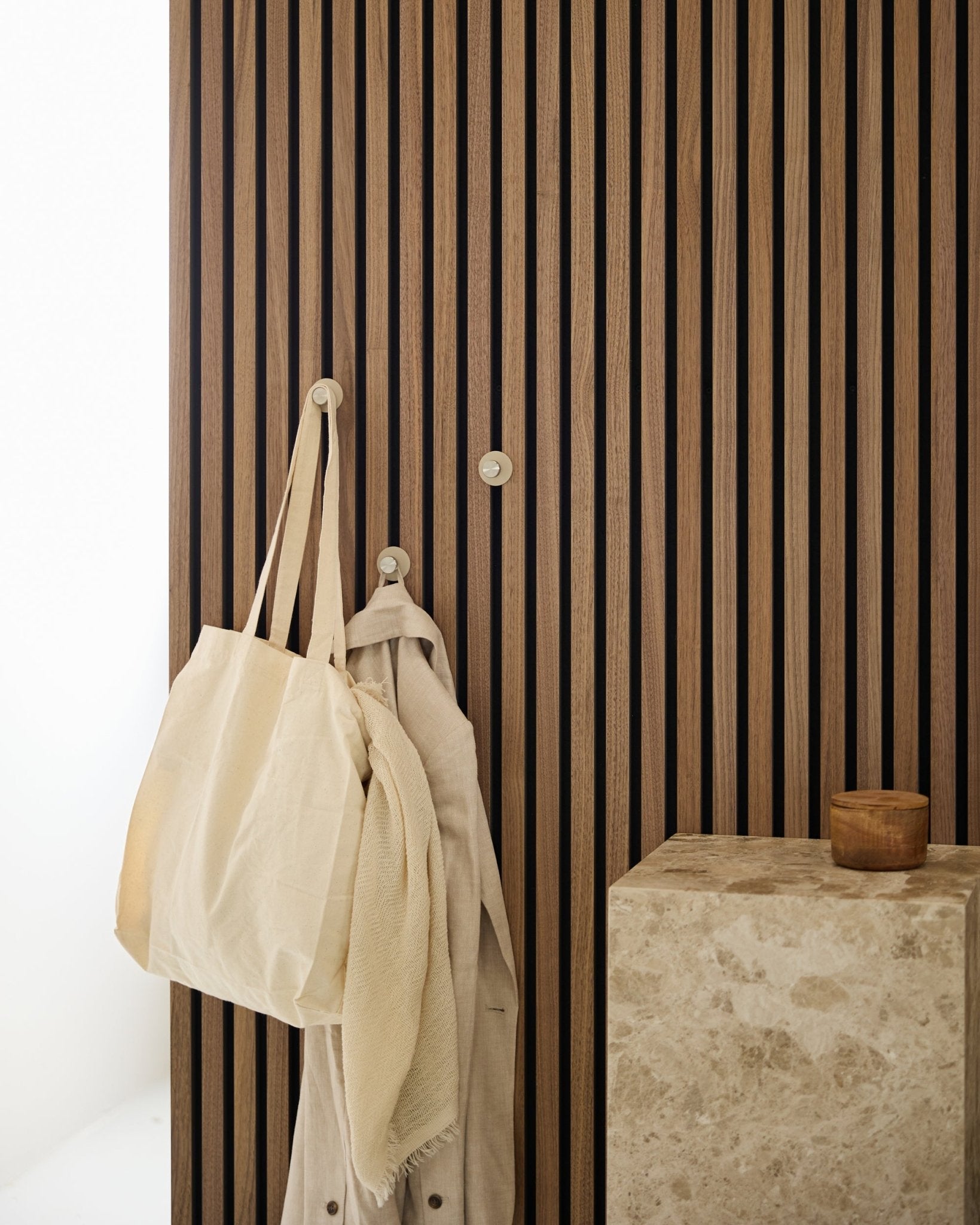 En beige tote bag og et lyst stykke stof hænger fra en WoodUpp Norr knage (Beige/rustfrit Steel), monteret på en vertical Akupanel Akustikpanel ved siden af en stenoverflade.