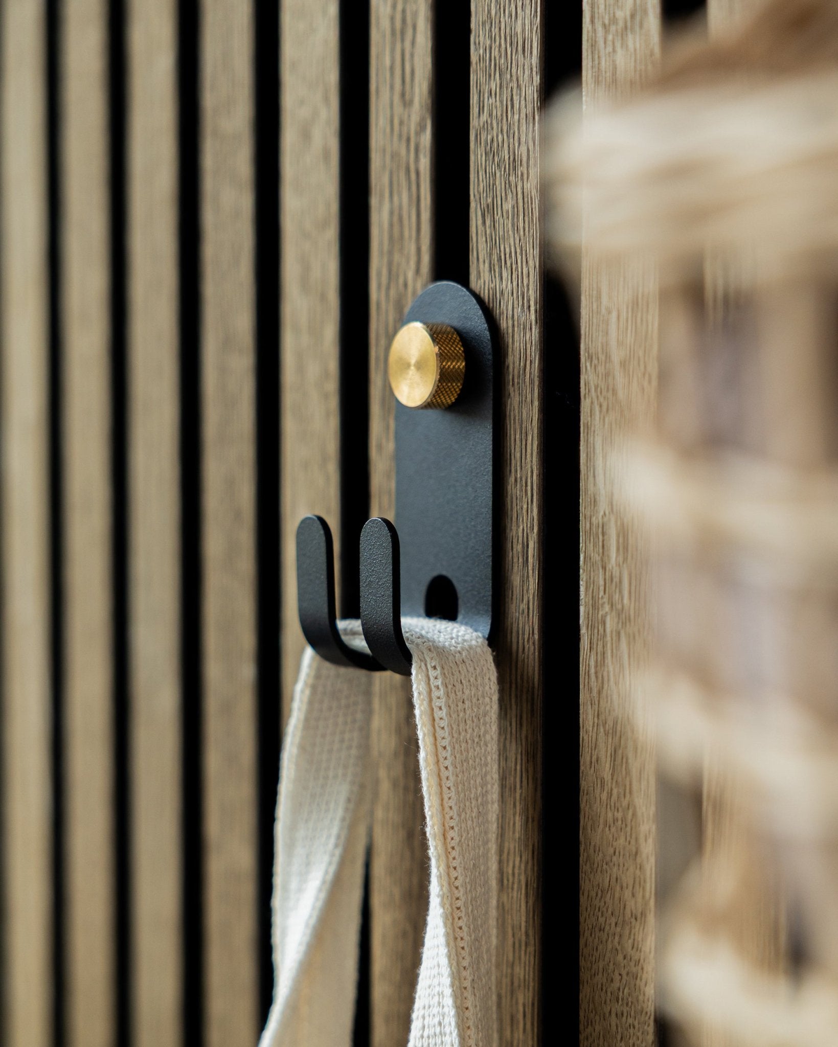 The WoodUpp Norr Double Hanger in black is mounted on a Nordic wooden acoustic slatwall panel, holding a cream-colored fabric strap.