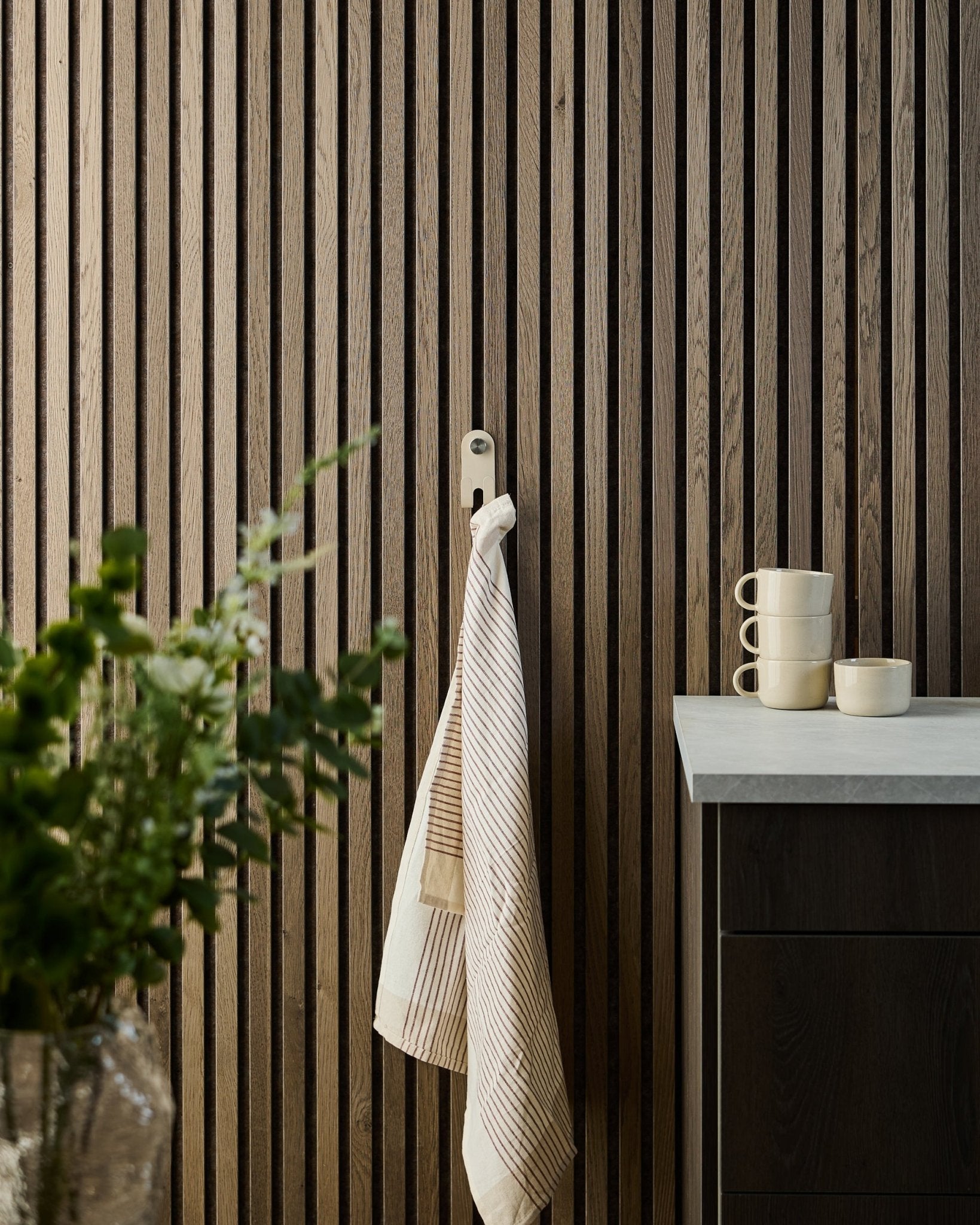 Et stribet håndklæde hænger på WoodUpp Norr Double Hanger i beige, monteret på et nordisk akustikpanel med lameller; krus, skål og en grøn plante står nedenunder.