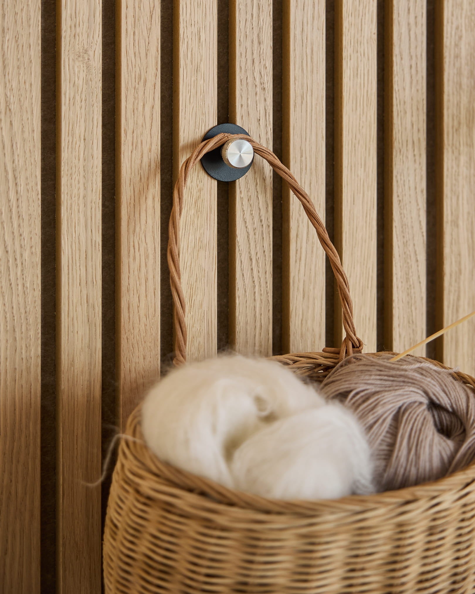 En kurv af hvidt og beige garn hænger på en Norr Coat Hook / X1, Black/Stainless Steel fra WoodUpp monteret på et Akupanel akustisk lamelvægspanel.