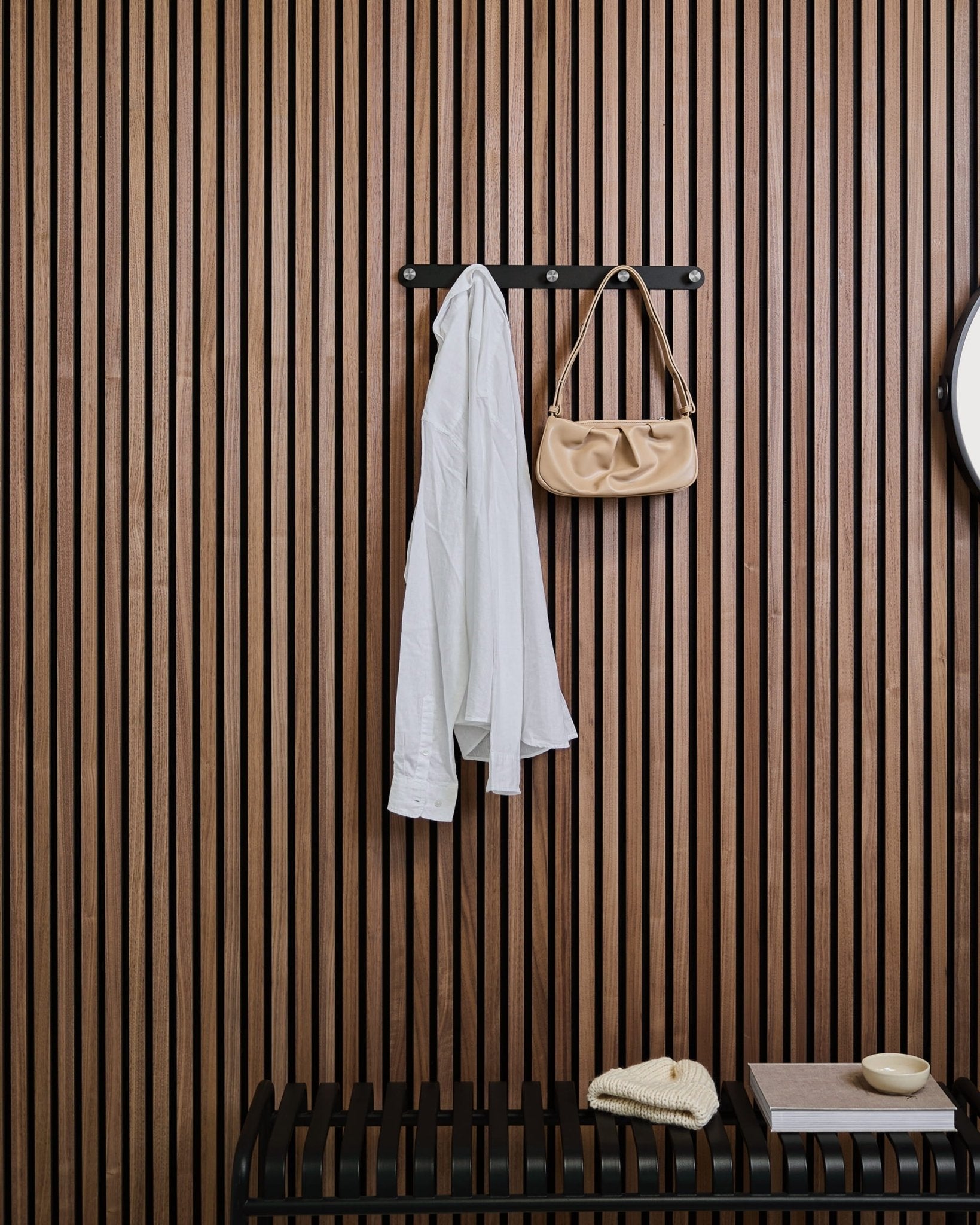En hvid skjorte og en beige håndtaske hænger på WoodUpps Norr Coat Hook Rack over en bænk mod en Akupanel-lamelvæg af træ med dekorationsgenstande.