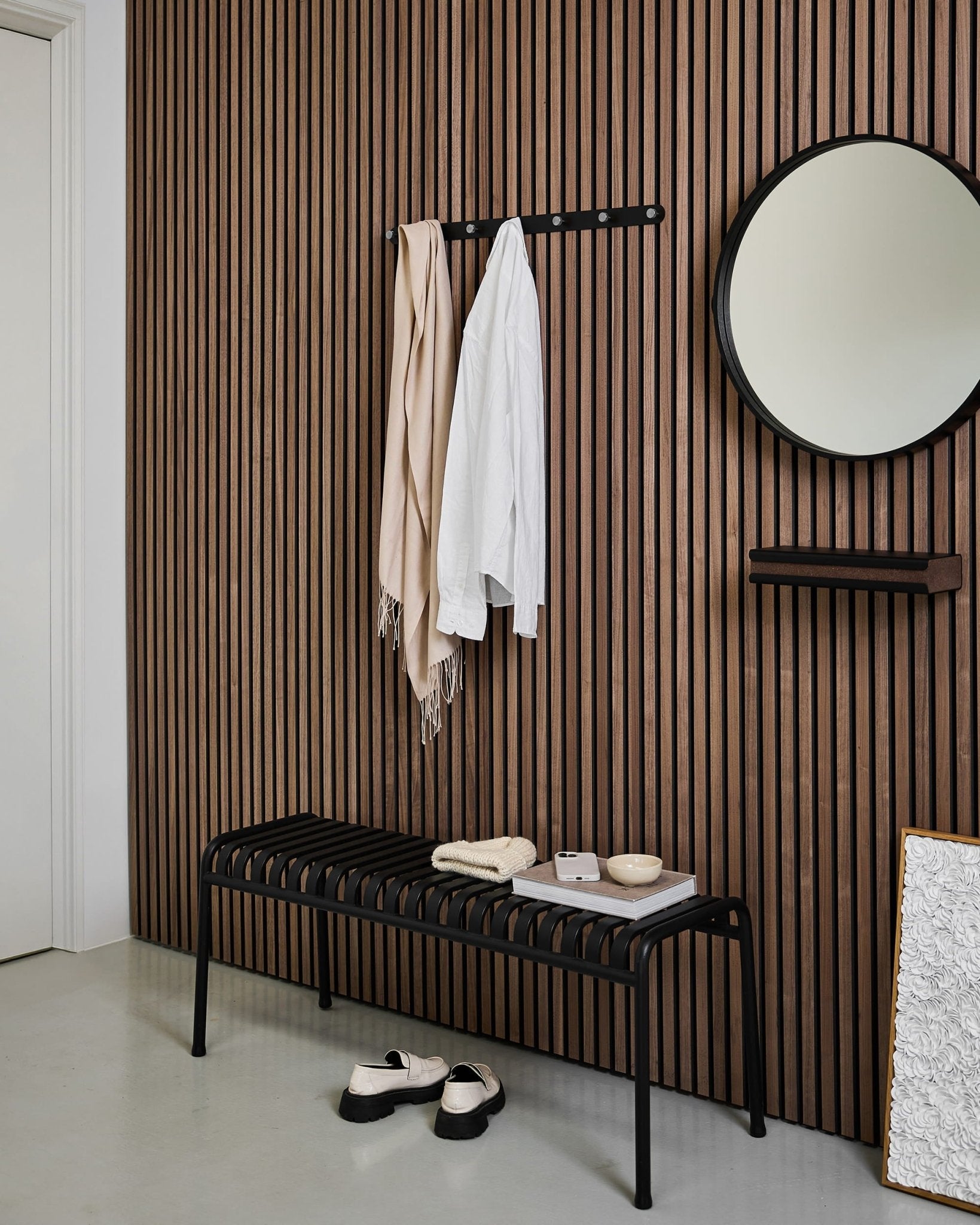 Moderne entré med en nordisk lamelvæg i akustisk træ, rundt spejl, sort bænk, WoodUpp Norr Coat Hook Rack og tøfler på et lyst gulv.